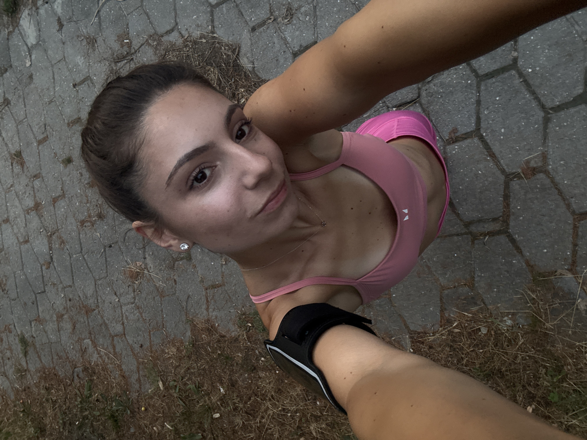 In Varna, Bulgaria, a young woman with dark hair pulled back in a bun takes a selfie outdoors. She is wearing a pink sports bra and pink leggings, with a black armband on her left arm. She has a delicate silver necklace on and diamond stud earrings. The ground is paved with gray cobblestones and dry grass.