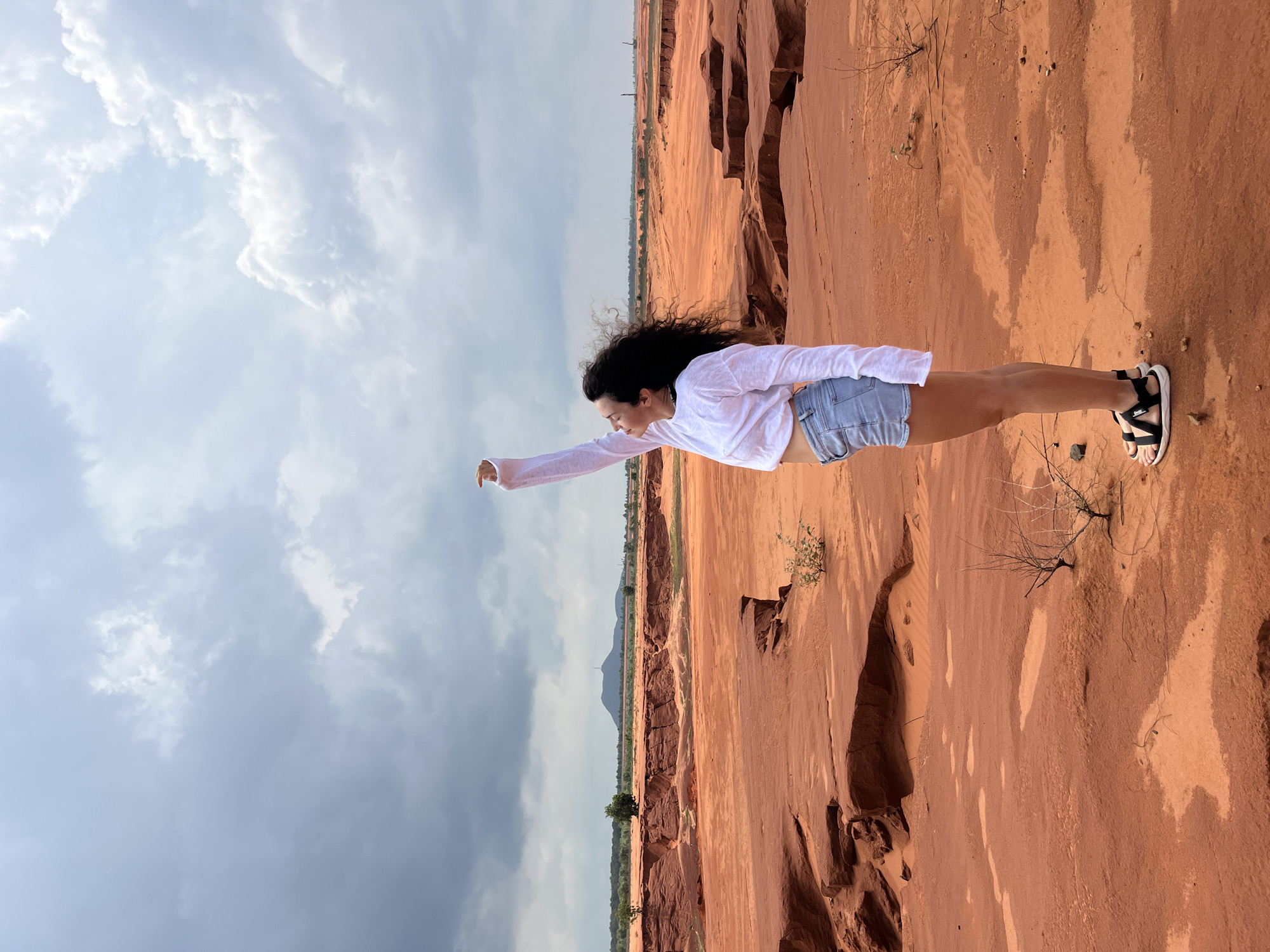 A woman stands in a desert-like landscape with red soil and eroded rock formations. She is wearing a white long-sleeved shirt and denim shorts, with her right arm extended upwards. The sky is cloudy. This scene is characteristic of the red sand dunes in Mui Ne, Vietnam.