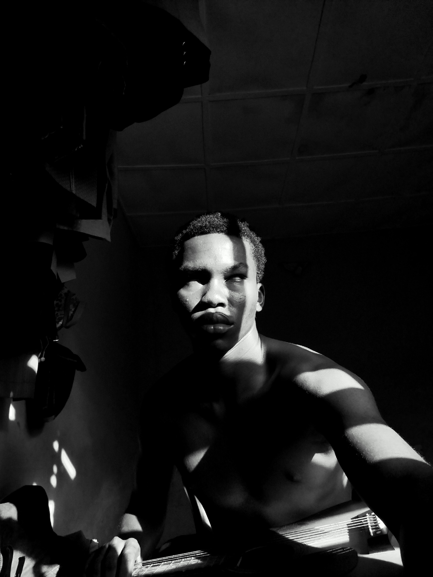 This black and white photograph captures a young man indoors, likely in Keffi, Nigeria, dramatically lit by natural light. The scene depicts a high-contrast environment with deep shadows and strong shafts of sunlight cutting through the darkness.
The central subject is a bare-chested young man with short, dark, curly hair. He is shown from the waist up, gazing upward and slightly to his right, with an expression that appears contemplative or focused. A prominent diagonal beam of sunlight bisects his face, illuminating his forehead, nose, and lips, while plunging the rest of his features into shadow. Similar stark patterns of light and shadow highlight the contours of his shoulders and chest.
His hands are visible in the lower right portion of the frame, gently holding the fretboard and strings of what appears to be a stringed instrument, likely a guitar. This suggests he is either playing or paused during a session with the instrument.
The background is largely obscured by darkness, with minimal details revealed. Above the man, a grid-like ceiling pattern is faintly visible, possibly indicating acoustic tiles or a similar structure. To his left, indistinct shapes, possibly clothing or objects, hang against a dark wall, catching glimmers of light. The strong directional sunlight indicates that the photo was taken during the daytime, likely when the sun was low in the sky, either morning or late afternoon. There is no visible text in the image.