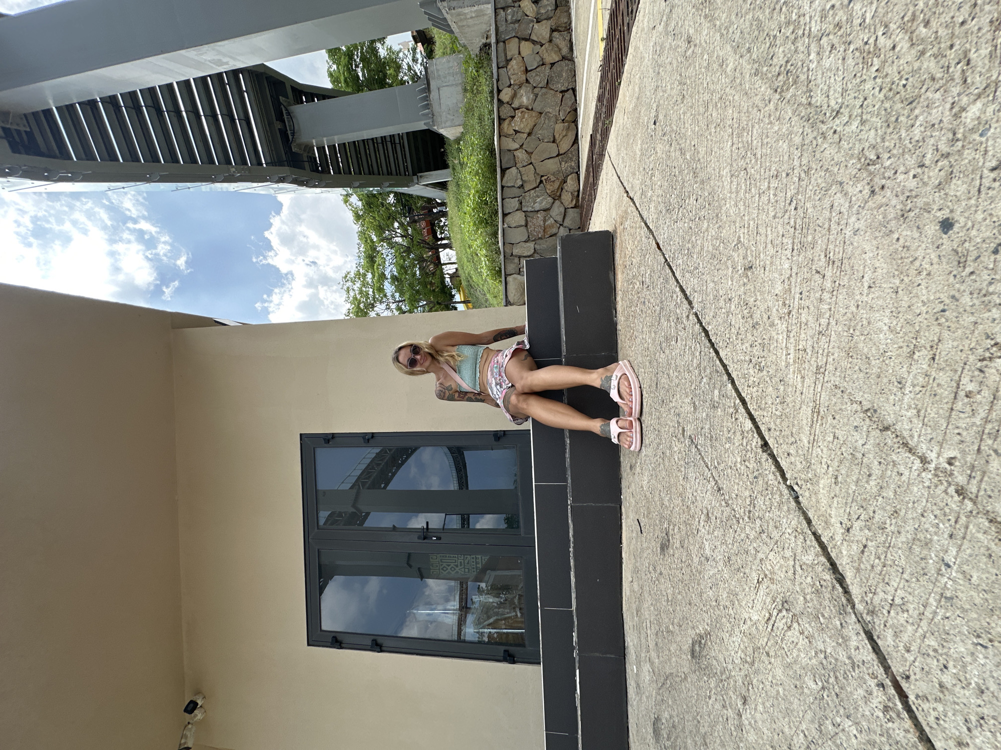 The scene depicts a light-skinned female individual seated on dark gray steps outside a light beige building under a bright, cloudy sky in Tien Thanh Ward, Vietnam. The individual has blonde hair, is wearing sunglasses, a light green crop top, a floral-patterned skirt, and pink flip-flops. Tattoos are visible on her left arm. She is positioned facing slightly towards the viewer, leaning against the building. Two white CCTV cameras are mounted on the upper left facade of the building. Dark gray double doors or windows are visible behind the individual, reflecting a large, elevated grey structural component, likely a bridge or overpass, and the sky. To the right of the building, a substantial grey concrete and metal elevated structure dominates the background, with the underside and supporting pillars visible. Below the structure and to the right of the steps, a grassy embankment is present, separated by a stone retaining wall. The foreground consists of textured concrete pavement with visible expansion joints and a metal drainage grate on the right. Trees and foliage are visible in the mid-ground.