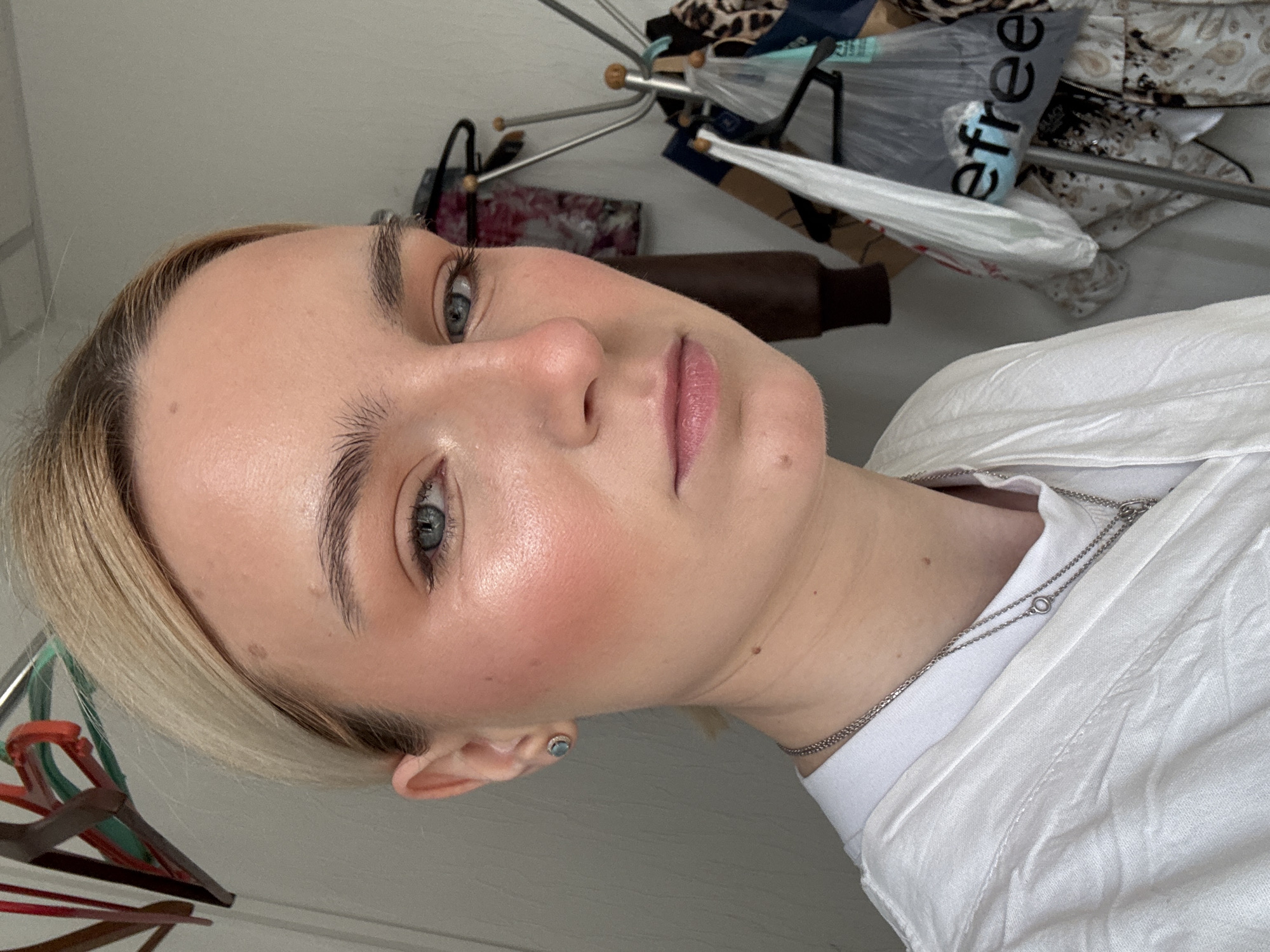 A close-up, tilted shot of a fair-skinned young woman with blonde hair and light blue eyes. She is facing slightly left of the camera. She is wearing a white shirt and a delicate silver necklace. An earring is visible in her left ear. The background contains a clothing rack with various articles of clothing and hangers. A shopping bag with the word "free" is also visible. The location cannot be determined from the information.