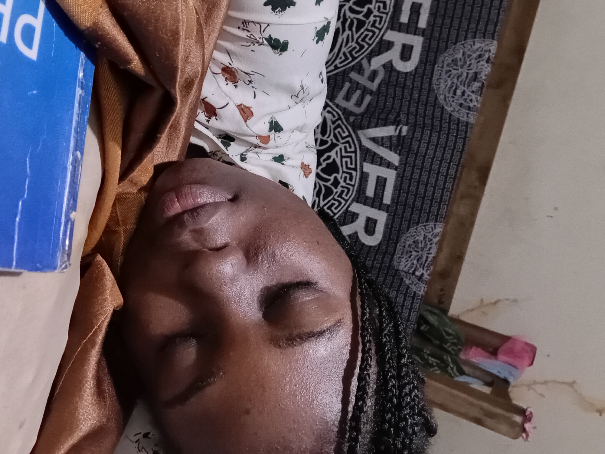 A close-up, high-angle shot shows a young Black woman with braided hair sleeping on a bed. Her eyes are closed, and her head is resting on a folded, golden-brown fabric. She is wearing a white shirt with a floral pattern. Behind her, a dark fabric with a repeating pattern of the word "VER" and a circular emblem is visible. To the upper right, a wooden structure with some colorful clothing draped over it is partially in view, set against a light-colored wall. In the lower foreground, a blue object with white lettering, possibly a book or box, is partially visible. The overall scene suggests a casual, indoor setting, likely a bedroom in a home in Ejule, Nigeria.