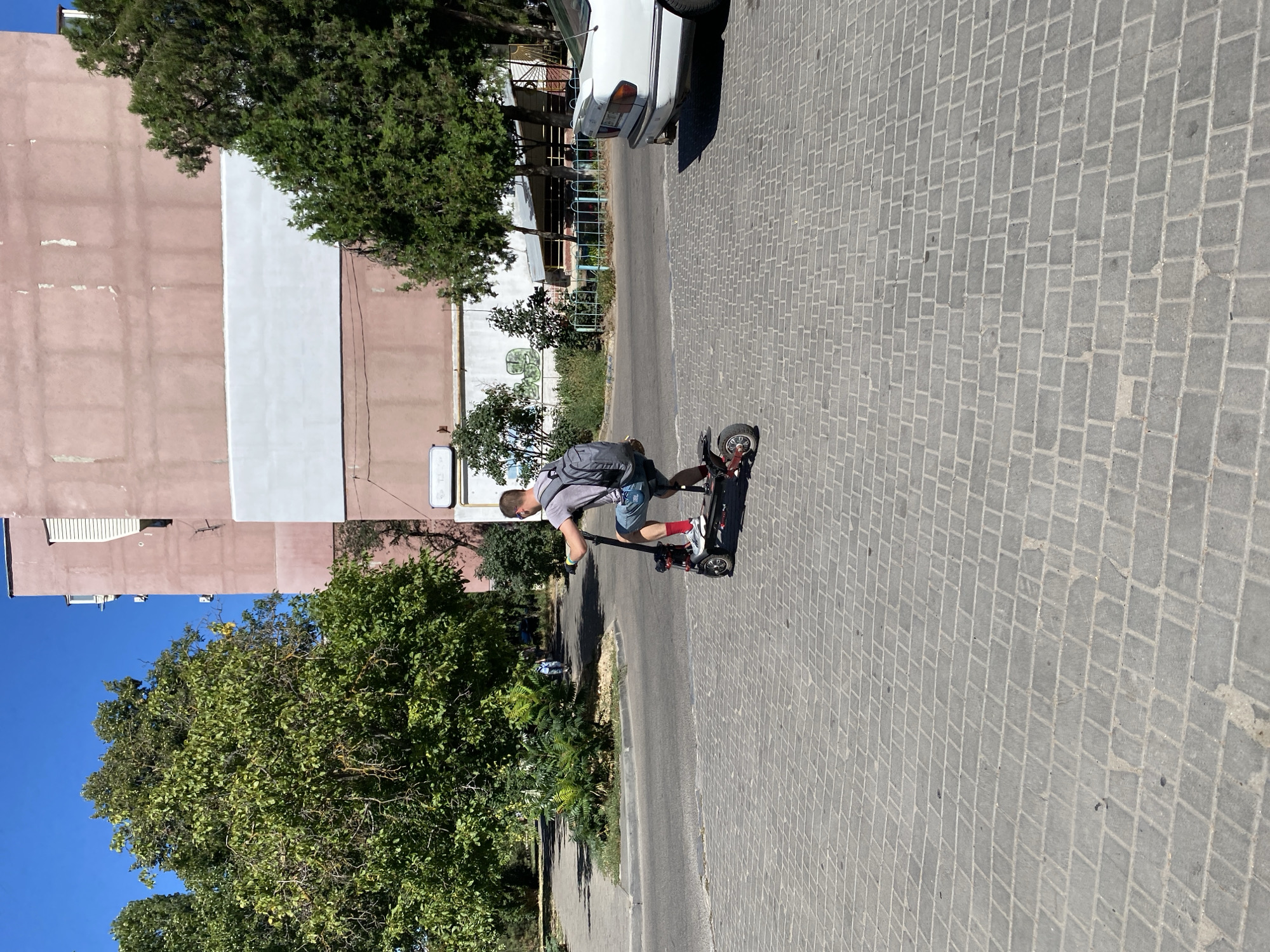 A young man wearing a grey t-shirt, blue shorts, and red socks is riding an electric scooter on a paved pedestrian walkway in Chornomorsk, Ukraine. He has a grey backpack on. The walkway is made of grey interlocking pavers. In the background, there is a multi-story building with pink and white sections, mature green trees, and a white car. The sky is clear and blue.