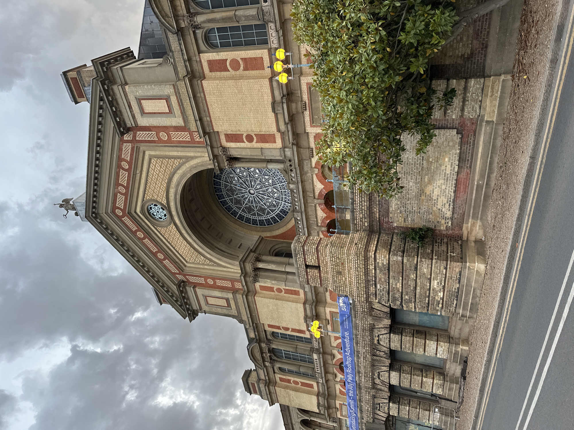 The image depicts a detailed view of the Alexandra Palace in London, England. The building is constructed with a combination of red and yellow bricks, and features a large, decorative glass window in the upper section. Architectural details include arches, columns, and intricate patterns. A tree partially obscures the view on the right side of the building. A street with white lines is visible in the foreground. There is an advertisement banner hanging on the side of the building. The sky is overcast with gray clouds.
