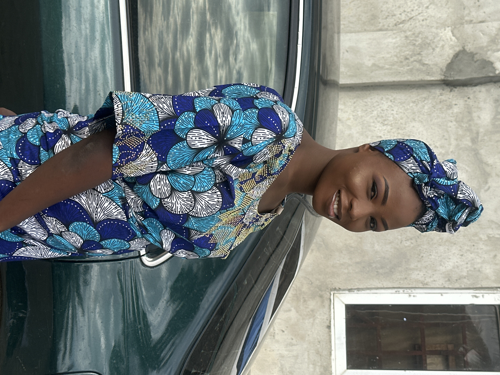 A young Black woman with a warm smile is pictured outdoors in Badagry, Nigeria. She is wearing a colorful blue and white patterned dress with a matching headscarf. Her skin is dark, and she has on makeup, including eyeliner and eyeshadow. The woman is leaning against what appears to be the side of a dark green car, the hood of which is visible in the foreground. Behind her, a grey concrete wall with a small window can be seen. The lighting suggests it is daytime, possibly late afternoon due to the presence of shadows. The overall mood of the image is cheerful and relaxed, captured in a candid moment. There is no visible text in the image.