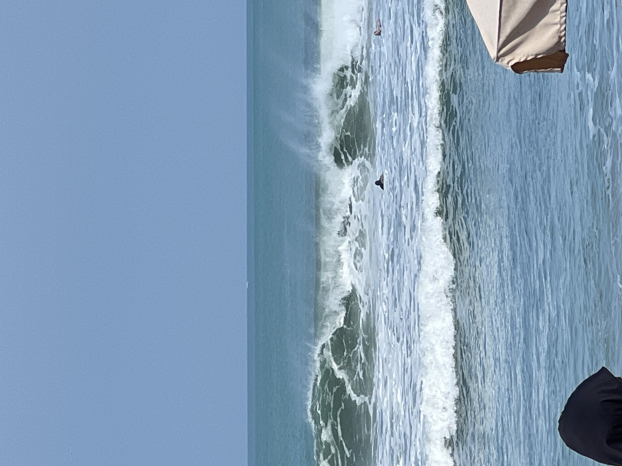 The image depicts a wide shot of the ocean at Kuta, Indonesia, on a bright, clear day. The sky is a pale blue, transitioning to a deeper teal where it meets the sea. The ocean is active, with significant waves breaking and churning white foam.
In the foreground, partially visible, is the side of a beige beach umbrella. To the far left, the top of a person's head, covered in a black garment, is visible.
Further out in the water, a surfer is visible in the middle ground, lying on their board and facing the shore. To the right of this surfer, another person is also in the water, possibly a swimmer or another surfer, though less clearly defined. In the far distance, a faint line suggests a boat or distant land on the horizon. The overall scene captures a typical beach day with moderate surf activity.