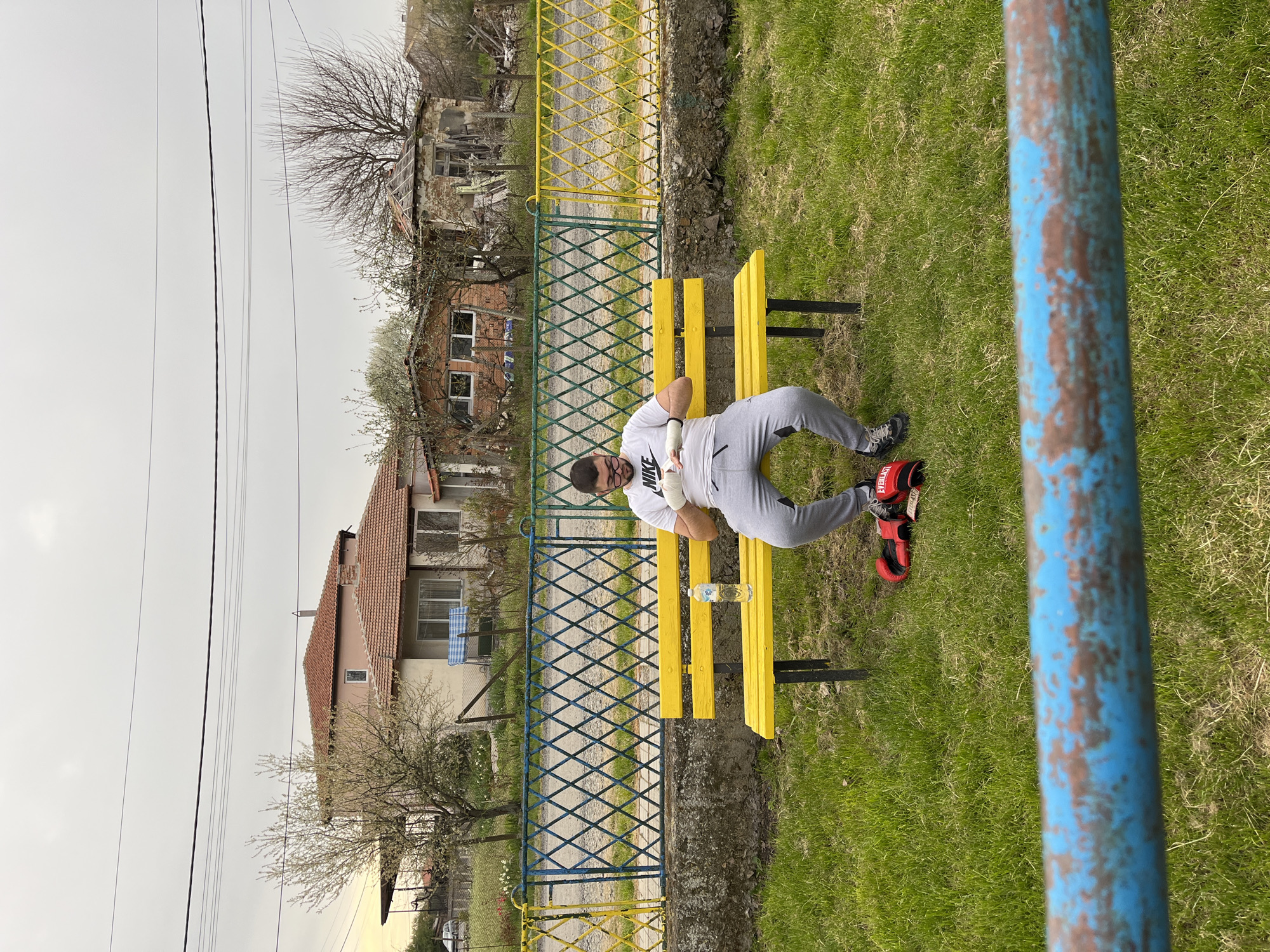 A horizontal, medium shot depicts a man resting on a yellow bench in Polski Izvor, Bulgaria. The man, wearing a white Nike t-shirt and grey sweatpants, is seated with red boxing gloves to his right and a clear water bottle positioned between him and the bench's center. A blue and yellow diamond-patterned fence spans the length of the background, behind which sits two residential houses of varying states of repair. Patches of green grass cover the ground in the foreground. A weathered blue pole is visible in the right foreground, slightly obscuring the view. The overcast sky suggests a gloomy day.