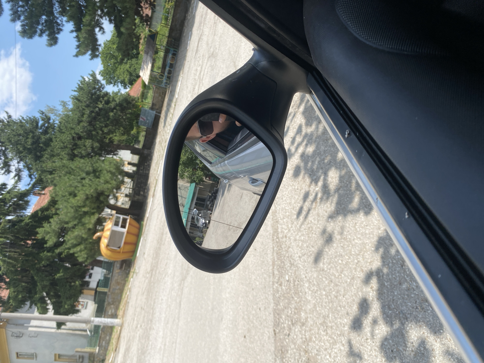 This is a close-up, slightly angled, eye-level shot of a car's side mirror, reflecting a scene in Hirevo, Bulgaria. The mirror itself is black and mounted on the car door. In the reflection, a person wearing sunglasses is visible through the car window, holding up a hand and appearing to take a photo. The background in the mirror shows a gray asphalt area, a building with a green roof, trees, and a unique, bright orange pumpkin-shaped structure with windows, likely a small kiosk or shop. Beyond that, there is more greenery and a bright blue sky with white clouds. The foreground of the image, outside the mirror, shows a portion of the car's interior, including the dark-colored door panel and a sliver of the car's exterior frame. The ground outside the car is a light-colored, textured asphalt, with shadows of trees cast upon it.
