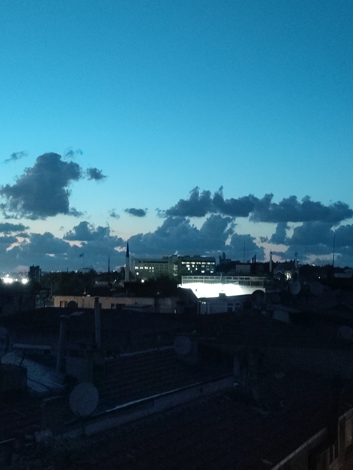 The image captures a cityscape at dusk, viewed from a high vantage point looking towards the horizon. The sky is a deep, clear blue, transitioning to a lighter shade near the horizon, with scattered dark clouds visible. Silhouetted against the sky are the rooftops of various buildings, predominantly dark and appearing to be residential or commercial structures. In the middle ground, a few taller buildings stand out. One appears to be a school or hospital building, brightly lit with rows of windows. To its right, a minaret of a mosque rises, illuminated from below. Another building with a distinctive pointed spire is also visible. The foreground is dominated by the dark, shadowed rooftops, some of which have satellite dishes. The scene is devoid of any visible people or vehicles. The overall impression is a calm, urban landscape at the end of the day, likely in Kadıköy, Turkey, given the architectural style.