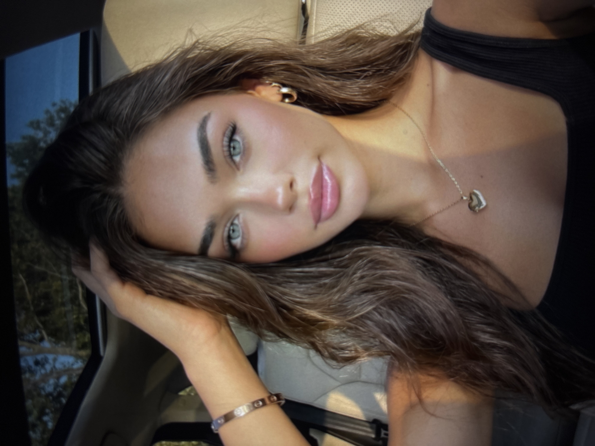 A young woman with long, wavy brown hair and bright blue eyes is pictured from the chest up, looking directly at the camera. She has fair skin, full lips with light pink lipstick, and prominent eyelashes. She is wearing a black tank top, a gold heart-shaped locket necklace, and hoop earrings. A silver bracelet with small embellishments is visible on her left wrist. Her left hand is raised and touching her hair, near the sunroof of a vehicle. The background shows the interior of a car with beige seats and a glimpse of trees and sky through the sunroof. The lighting suggests it is daytime, possibly late afternoon due to the warm, golden hue. The woman appears to be in a car, likely in or near Kyustendil, Bulgaria, though no specific landmarks confirm this location.