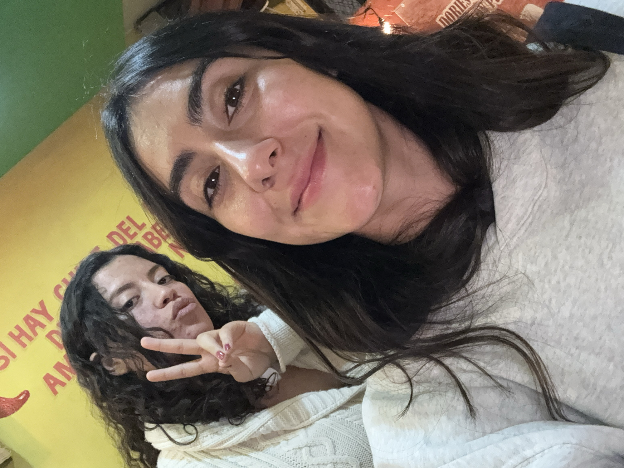 Two females are depicted in a selfie taken indoors in San Bernardino Tlaxcalancingo, Mexico. The foreground subject, positioned off-center to the right, has long, dark, wavy hair and a subtle smile, wearing a light gray long-sleeved top. Her face is well-lit, showing natural skin tone. The background subject, behind and to the left of the foreground subject, has dark, curly hair and is wearing a white cable-knit cardigan. She is looking at the camera with a playful expression, her lips slightly pursed, and displays a "peace sign" gesture with her right hand, featuring dark pink nail polish. The background immediately behind her consists of a bright yellow wall adorned with a red chili pepper graphic and red text, partially visible as "SI HAY AM... DEL... RE...". To the right of the foreground subject, a blurred, warm-toned orange or red surface is visible, showing partial text that reads "DORIES". A small segment of a green ceiling or wall is visible at the very top of the frame. The lighting suggests an indoor setting with artificial illumination.