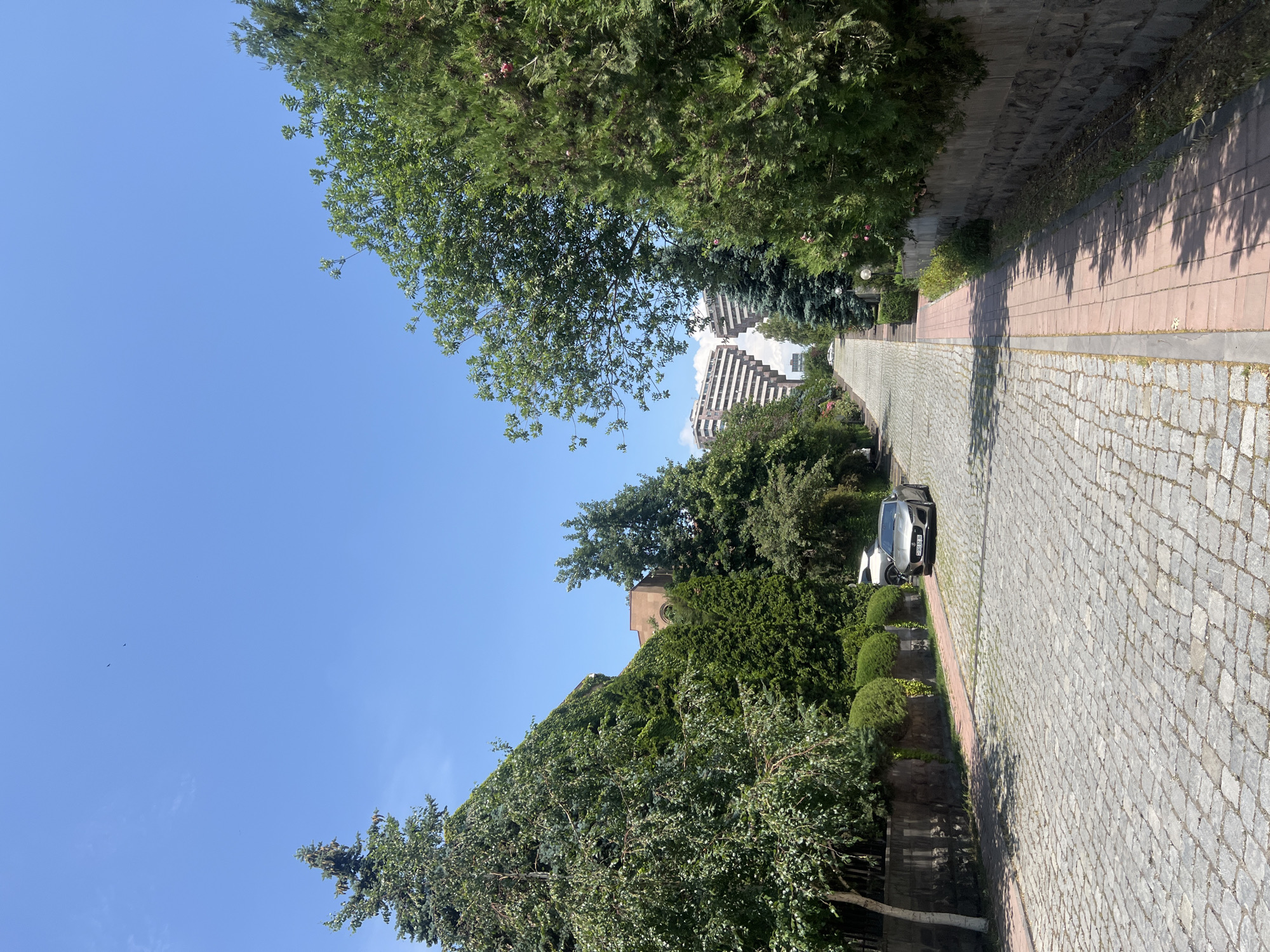 The image shows a cobblestone street in Yerevan, Armenia, leading uphill towards a distinctive architectural complex. The street is lined with lush green trees and hedges on both sides, with stone walls bordering the properties. A modern silver car is parked on the street, and a pedestrian walkway made of red tiles runs along the right side. In the distance, two white buildings with unique stepped facades, reminiscent of pyramids, are visible against a clear blue sky. The overall impression is one of a well-maintained urban street with architectural landmarks.