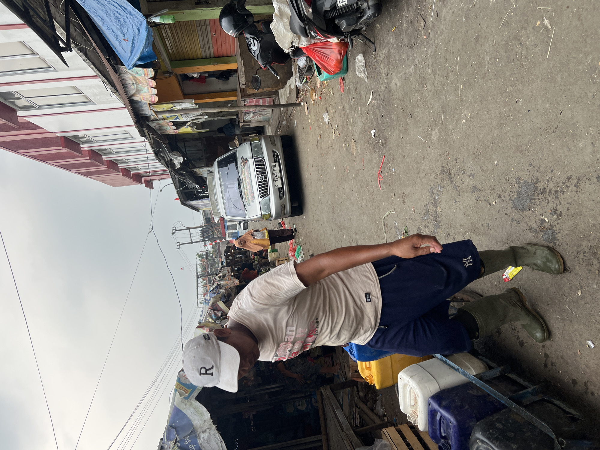 In Citeureup, Indonesia, a man wearing a white cap with an "R" logo, a light-colored t-shirt with graphic print, dark blue shorts, and green rubber boots is walking down a dirt path lined with market stalls. He is positioned in the foreground, looking downwards and to his left. To his left, a cart holds several large plastic containers, one yellow and one dark blue, filled with liquid. In the background, a silver van with the license plate "F 1745 FNL" is parked. Further back, a woman in a headscarf and yellow shirt is visible. The market stalls feature various goods, and the ground is littered with debris, including plastic bags and wrappers. A black motorcycle is parked on the right side of the image, and the scene appears to be under an overcast sky.