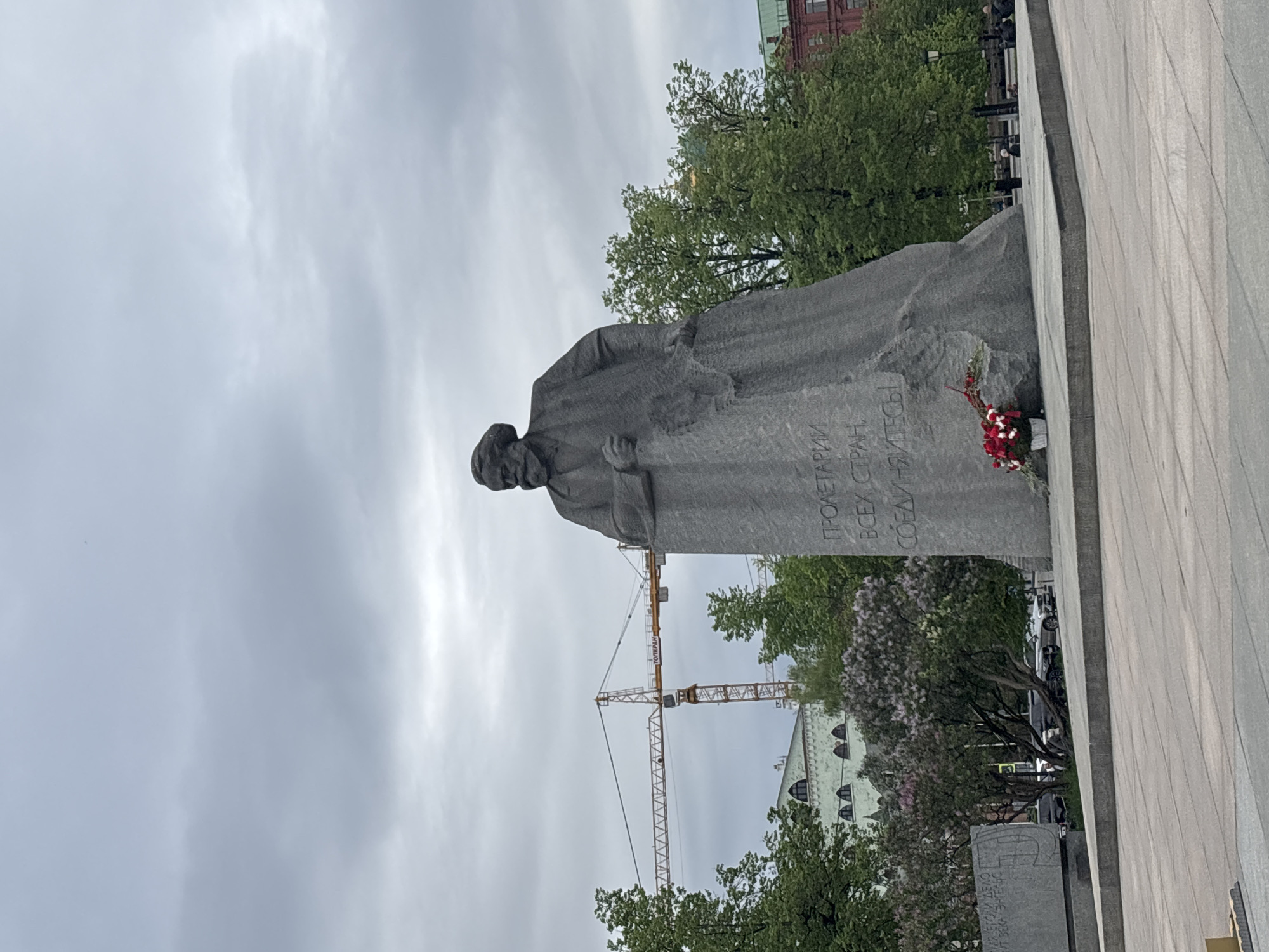 The image shows a statue of Karl Marx in Moscow, Russia. The statue is made of gray stone and depicts Marx in a long coat, looking to his left. The inscription on the statue is in Russian. At the base of the statue is a wreath of flowers. In the background, there is a construction crane, trees, a building with arched windows, and a cloudy sky. The statue is placed on a grey stone surface.