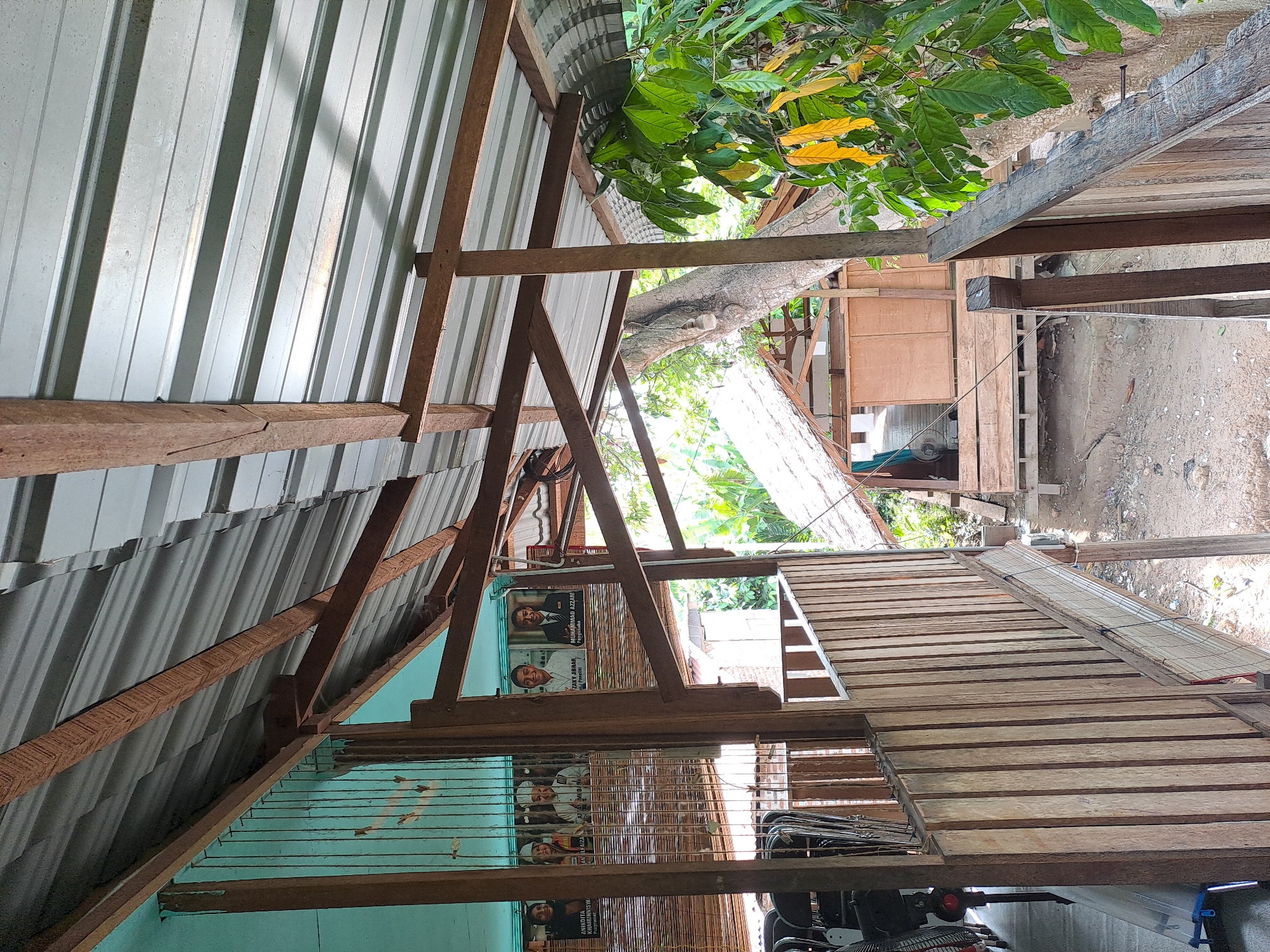The image depicts a rustic, open-air structure with a corrugated metal roof supported by wooden beams, located in Geulanggang Baro, Indonesia. The scene suggests a partly built or maintained area. In the background, a small wooden building with a thatched roof is visible amidst lush green foliage and a large tree. Several framed portraits, seemingly of individuals in formal attire, are displayed on a light blue wall. Wooden railings and a pathway made of wooden planks are prominent features, indicating a raised walkway or balcony. The ground below appears to be unpaved. The overall impression is one of a rural or semi-rural setting, possibly a community center or a residential area with open-air extensions.