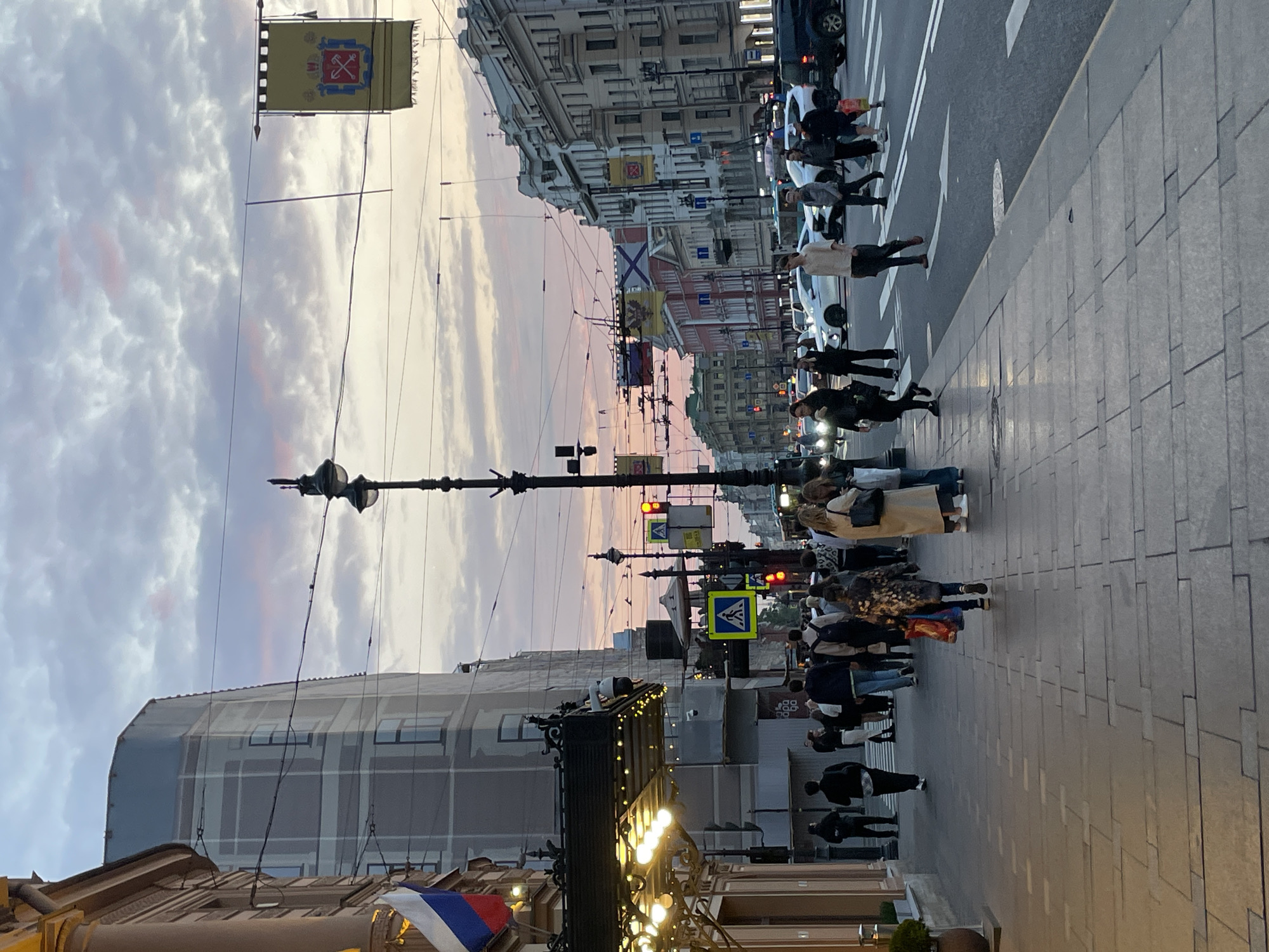 The image depicts a street scene in Saint Petersburg, Russia, likely Nevsky Prospekt. Several pedestrians are crossing the street at a crosswalk. Traffic lights are visible and appear to be red. Banners with the coat of arms of Saint Petersburg hang from overhead wires, while Russian flags are visible at the lower left. The buildings on either side of the street are multi-story, with classical architectural details. The sky is overcast with a pinkish hue, indicating either sunrise or sunset. A street lamp is visible in the foreground.