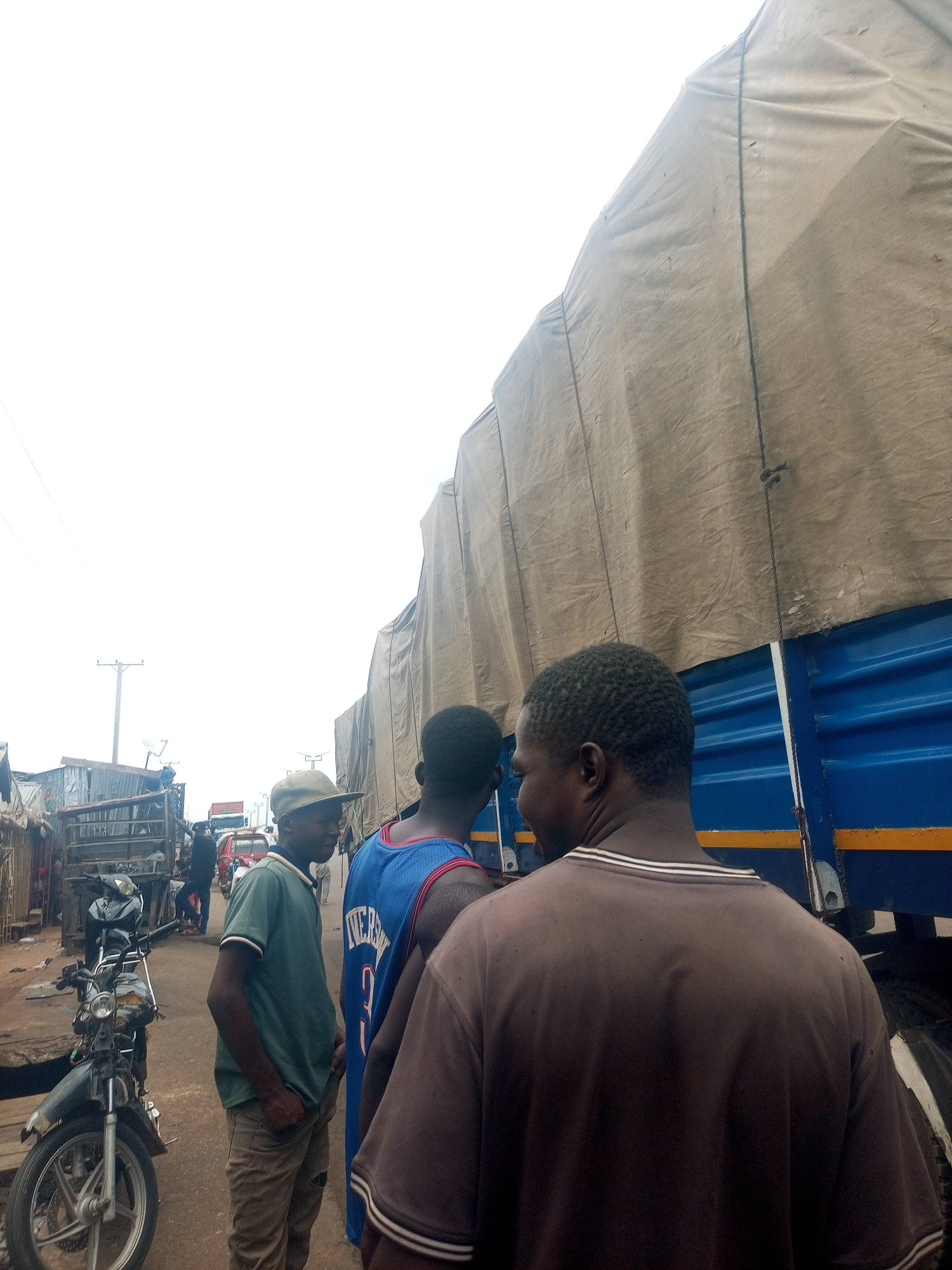 The image depicts three men standing near a large, blue truck with a tan-colored tarp covering its cargo in what appears to be a crowded street. A partially disassembled motorcycle is parked to the left. The man in the foreground is wearing a brown shirt and faces the truck. To his left, another man wears a blue basketball jersey. The third man, on the left, is wearing a green shirt and a baseball cap. The background includes other vehicles, buildings, and what appear to be utility poles. Based on the prompt information, the location is Gombe, Nigeria.