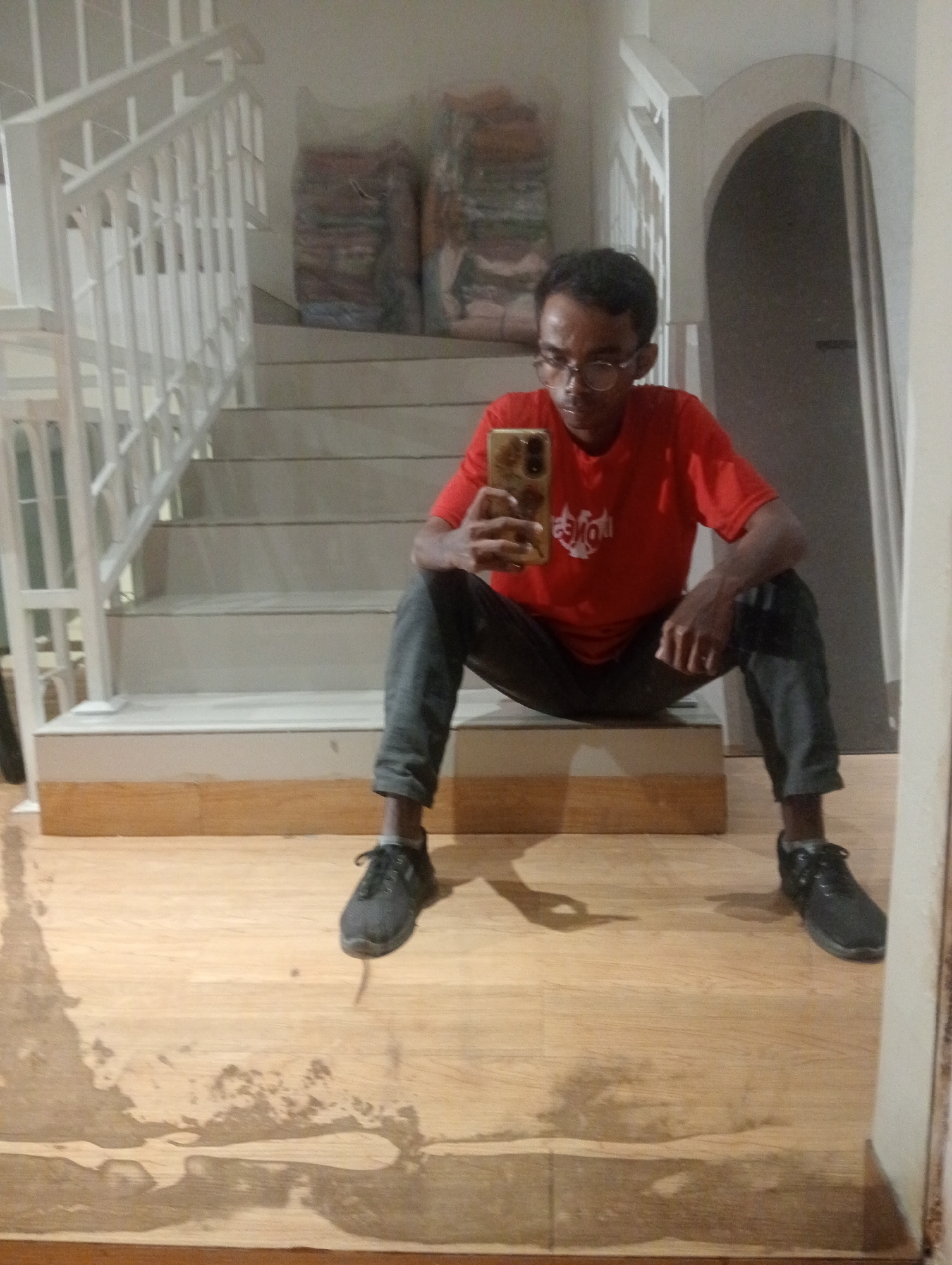 The image shows a young man with dark skin and short, dark hair, wearing glasses, a red t-shirt with a white logo, and dark grey pants. He is sitting on the bottom step of a white staircase, facing a mirror and taking a selfie with his phone, which has a patterned case. The staircase has white railings and appears to be indoors. Behind the staircase, there are some stacked items covered in plastic wrapping. The floor in front of the stairs is made of light-colored wood planks, which are reflective and show some water stains or patches. The overall lighting suggests it might be daytime or a well-lit indoor space, but there are no direct cues for time of day or weather. The man's expression is neutral as he looks at his phone. The scene appears to be a residential interior, possibly a hallway or entrance. The text on the man's t-shirt is visible but blurry, with white lettering that is difficult to decipher.