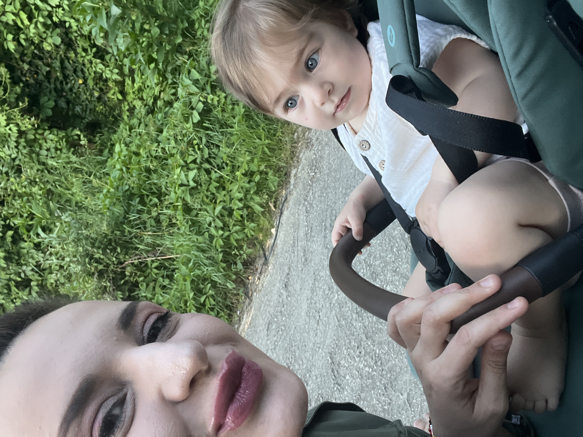 A close-up, selfie-style photograph captures an adult and a young child outdoors on a bright day. The adult's face is partially visible in the bottom left, featuring prominent dark pink, glossy lipstick, groomed eyebrows, and eye makeup. They are looking slightly upwards towards the camera.
To the right, a baby or toddler with light blonde hair and striking blue eyes sits in a green or teal-colored stroller, looking directly at the viewer with a neutral, curious expression. The child wears a white, textured top with light-colored buttons and is secured by black safety straps. Their bare legs and feet are visible within the stroller.
The adult's hand is seen holding the stroller's dark brown handlebar, with a small bracelet on their wrist. The background and left side of the image are filled with lush, vibrant green foliage and bushes, indicating a natural outdoor setting like a park or roadside. A paved, grey path runs through the scene, separating the green areas. The lighting suggests it is daytime, with ample natural light. No clear text is visible.
