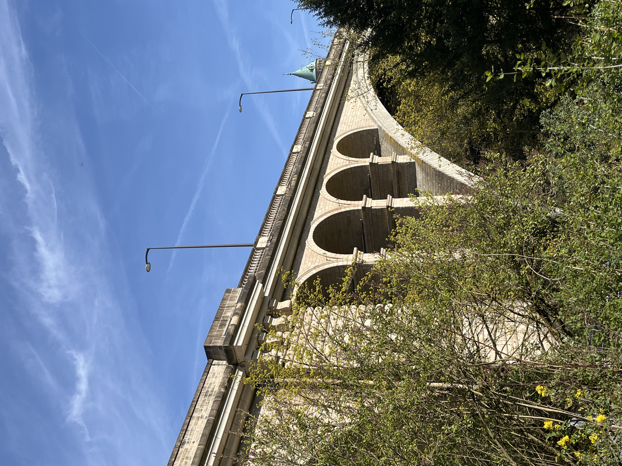 The image shows the Passerelle Viaduc in Luxembourg City, Luxembourg, viewed from a low angle. The viaduct, constructed of light-colored stone, features a series of arches supporting a walkway lined with a balustrade. Streetlights are positioned along the bridge's edge. A green spire is visible above the structure. The sky is blue with wispy clouds. Trees and foliage surround the base of the viaduct on the right side.