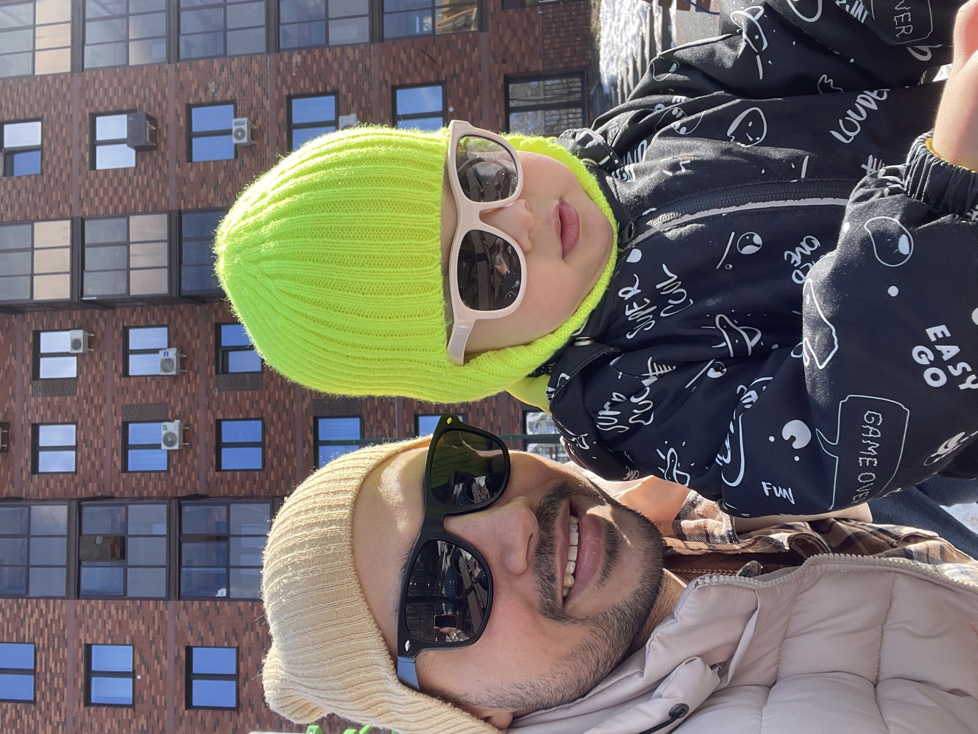 A selfie shows a man and child in front of a building in Izhevsk, Russia. The man, wearing a beige beanie and black sunglasses, smiles at the camera. A child, also facing the camera, wears a bright green beanie and light-framed sunglasses. They are in front of a brick building with many windows and window-mounted air conditioning units. The child is wearing a black jacket with white cartoon doodles. The man is wearing a tan quilted jacket over a plaid shirt. Icicles can be seen near the top of the photo.
