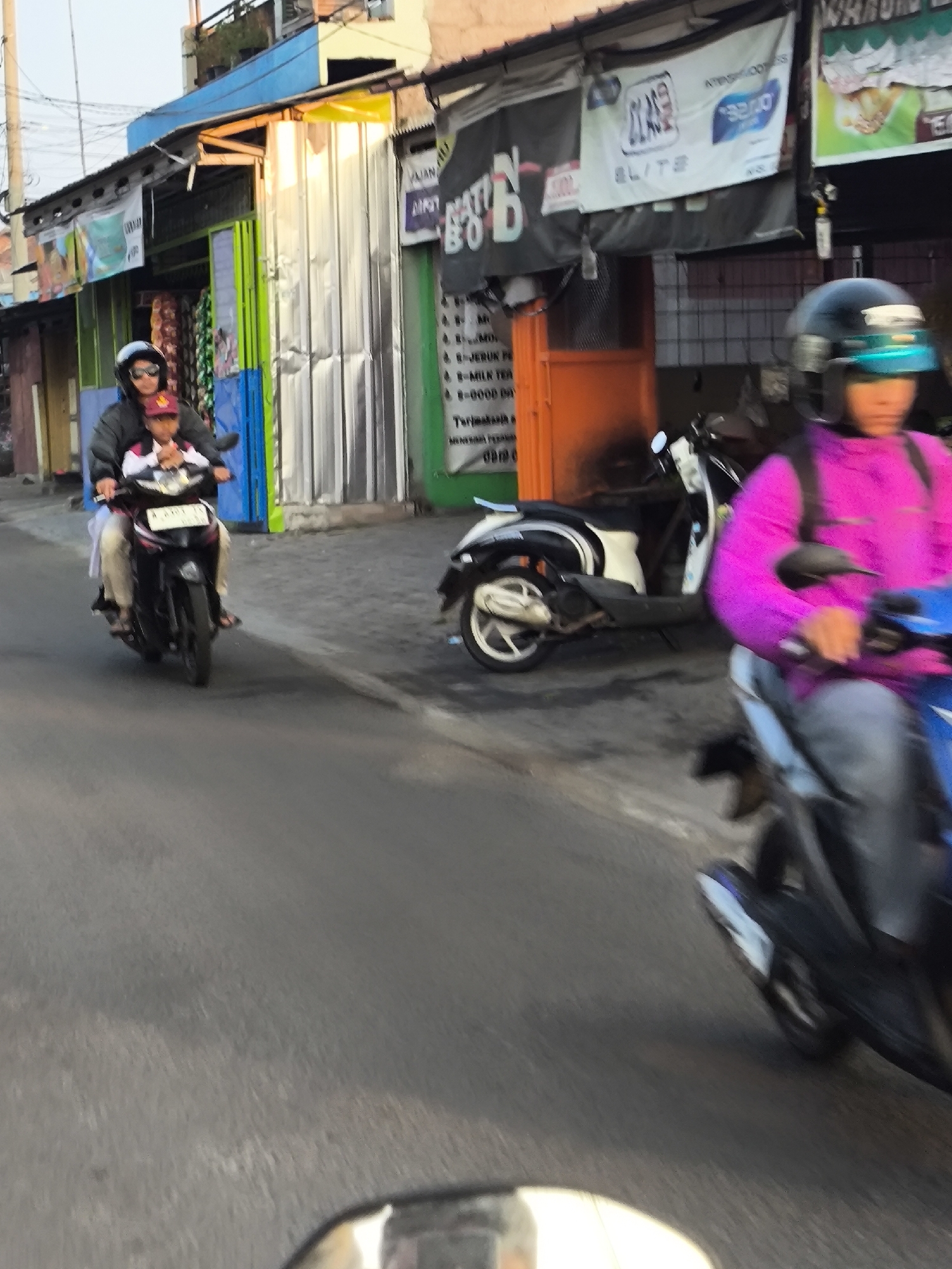 In Cilegon, Indonesia, two individuals are captured on a motorcycle on a street. The rider, wearing a dark jacket and sunglasses, is in front, with a child seated behind them, wearing a red cap and a white shirt. They are riding a black scooter with a license plate. In the background, a store with a yellow awning and silver corrugated metal walls is visible, displaying various banners and advertisements. To the right, another person on a motorcycle is seen wearing a bright pink jacket and a helmet with a blue visor, also in motion. The scene suggests a typical street activity in a commercial area of the city.