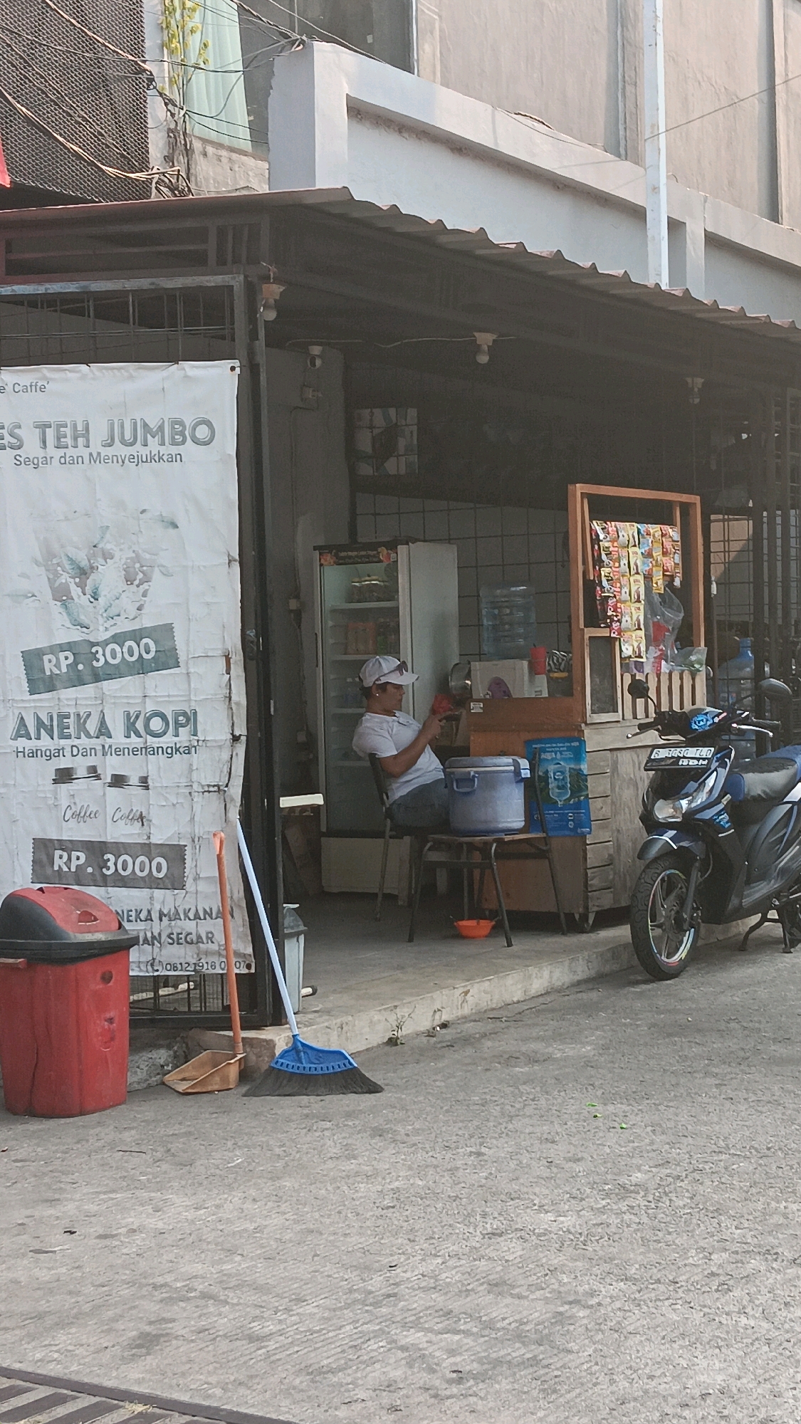 The image depicts a street-level view of a small shop in South Jakarta, Indonesia. The shop appears to be an outdoor stall with an awning providing shade. A woman, wearing a white t-shirt and a white cap, is seated on a stool, looking at her phone. She is positioned in front of a cooler and a stack of wooden crates. Various items, including bottled water, snacks in packaging, and what appears to be a menu or advertisement, are displayed on shelves behind her. A blue motorcycle is parked to the right of the shop, with its license plate visible. In the foreground, a red trash bin, a dustpan, and a blue broom are placed on the ground. A banner on the left side of the image advertises "ES TEH JUMBO" (Jumbo Iced Tea) and "ANEKA KOPI" (Assorted Coffee), with prices listed as Rp. 3000. The ground is paved with concrete. The overall scene suggests a casual, everyday setting in an urban environment.