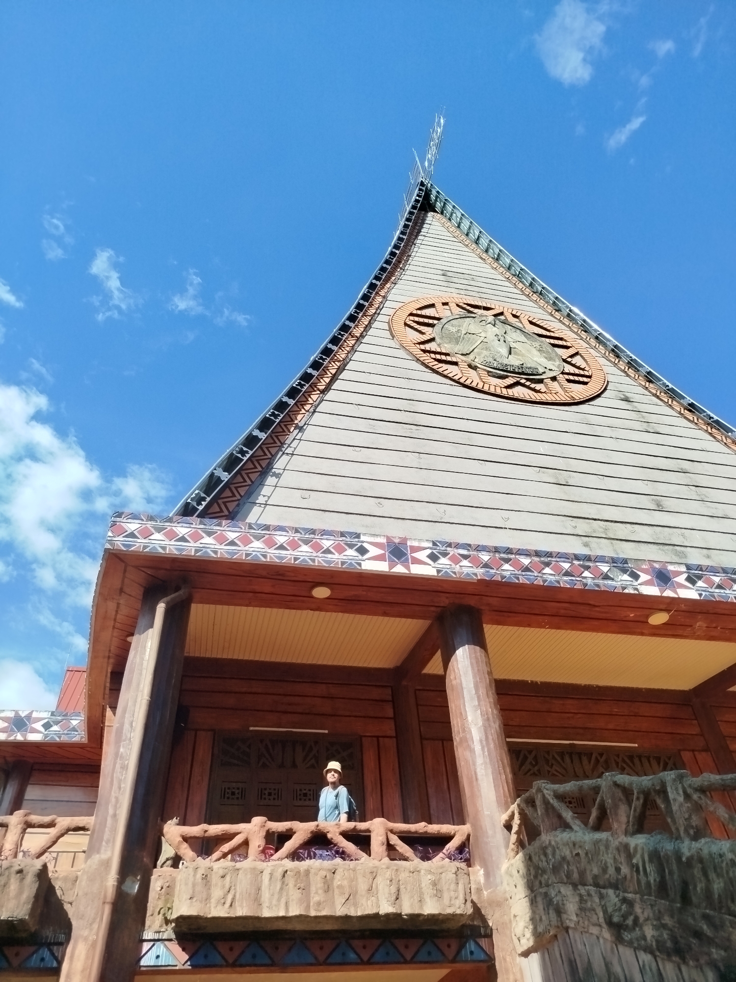 A low-angle, eye-level shot reveals a large wooden building under a bright blue sky with scattered clouds. The building's roof is steeply pitched and triangular, with a circular emblem near the peak. The emblem appears to be a carving or relief. The building's design incorporates intricate geometric patterns along the eaves. Below the roof, a balcony with a textured, faux-stone railing is visible. A single person wearing a hat and backpack stands on the balcony. The building's architectural style suggests a cultural or religious function. The image was taken in An Phu Ward, Vietnam.