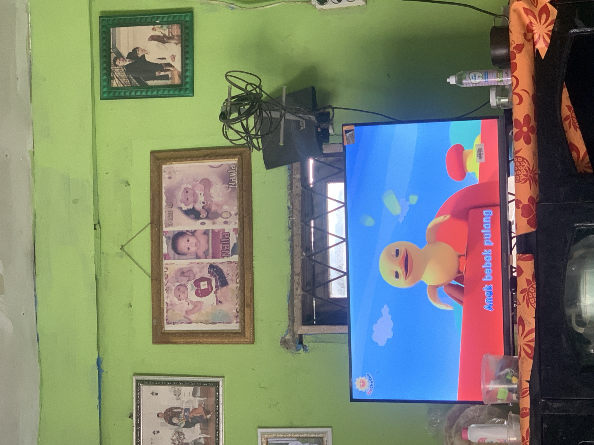 The image shows a television screen displaying a cartoon duck character on a bright blue background. Below the character, text reads "Anak bebek pulang". The TV is placed on a dark wooden cabinet covered with an orange patterned cloth. On the cloth, to the left of the TV, is a white plastic bottle with a pink cap, and a small clear container. To the right of the TV are two small jars, a dark mug, and a green bottle labeled "MY BABY". The wall behind the TV is painted light green. Several framed pictures hang on the wall. On the left, a framed photo shows a family. In the center, a framed picture displays two babies, with the names "Naila" visible. On the right, a framed photo depicts a couple. Above the TV, a window with a wooden frame is visible, showing a dark interior. The overall scene appears to be in a home environment, possibly a living room. The specific location in Palembang, Indonesia, cannot be determined from the image.