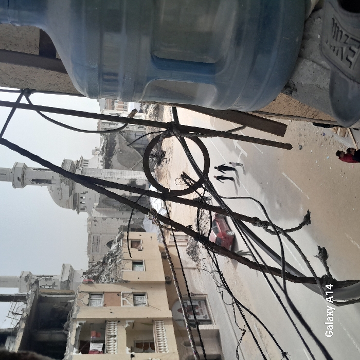 The image depicts a street scene in Khan Yunis, Palestinian Territory, characterized by significant structural damage. In the foreground, viewed from an elevated perspective, a large, translucent blue plastic water bottle occupies the right side, partially obscuring a dark sandal below it. Intricate, rusty metalwork, possibly part of a railing or security bars, extends diagonally across the frame from the upper left to the lower center-right, with multiple tangled wires attached.
Below the metalwork, at street level, a paved road shows patches of dirt and debris. Three individuals, two adults and a child, walk in silhouette towards the left side of the frame on the road. Further down the street to the left, a red sedan is parked facing away from the viewer. Along the left side of the street, multi-story residential buildings are visible, many exhibiting severe structural damage, with sections of walls and roofs collapsed, exposing interiors and twisted rebar. Clothes hang from balconies on an intact section of one building. Piles of rubble and debris are scattered along the roadside and in open areas.
In the background, a prominent mosque with a large dome and two tall minarets stands amidst further destruction. The mosque itself appears heavily damaged, with large sections missing from its structure, particularly on the lower levels and around the dome. The sky is a pale, hazy white, indicative of an overcast or dusty day. In the bottom right corner, partially obscured by the metalwork, two more individuals, one in a red shirt, are visible at a lower vantage point.