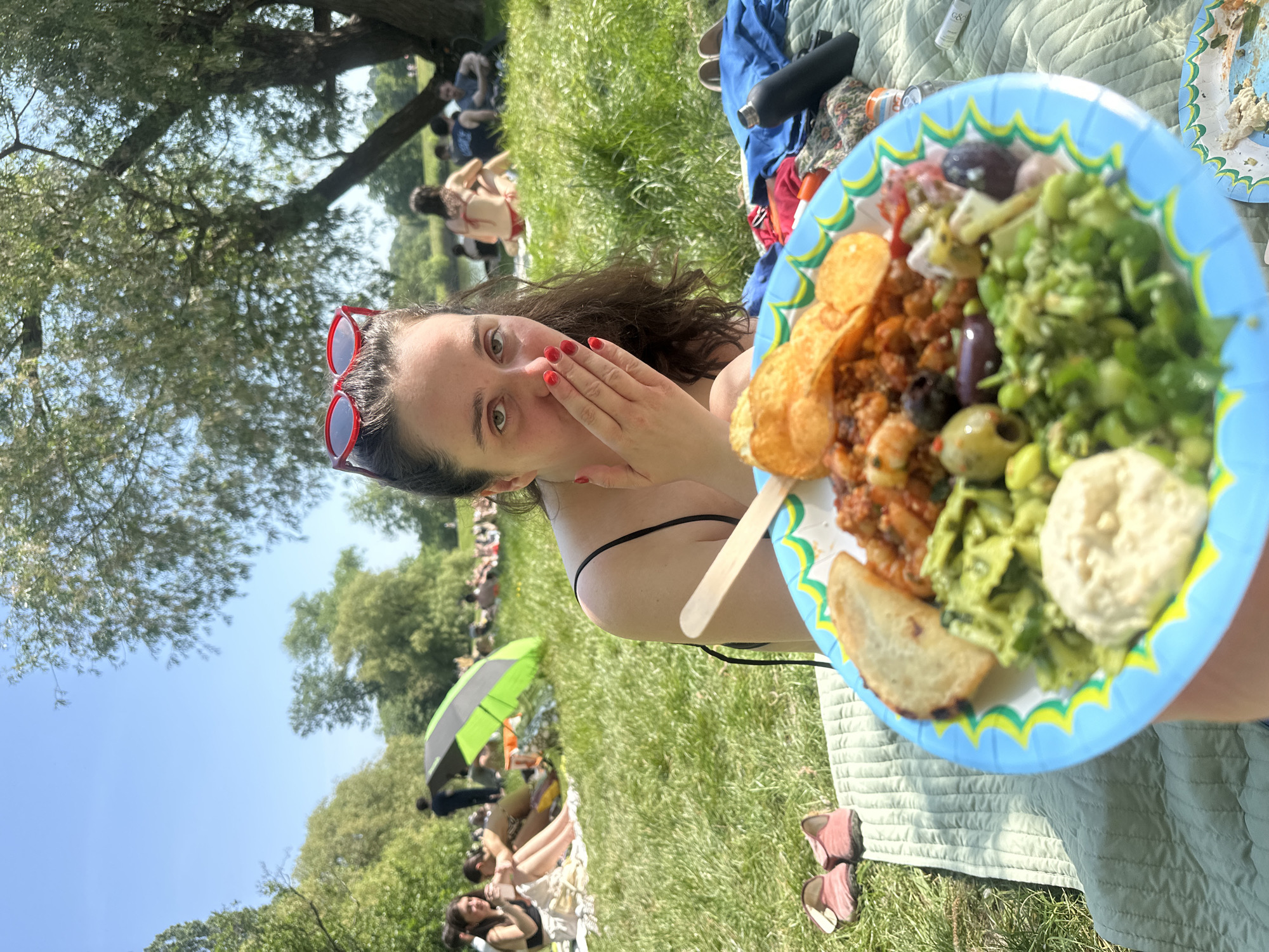 The image shows a woman holding a paper plate of food, seemingly enjoying a picnic in a grassy area. The plate contains a variety of items, including chips, olives, pasta, and what appears to be hummus. In the background, other individuals are relaxing on the grass, some under an umbrella. Given the style of greenery, weather and the attire of people, the scene is likely taking place in a park in London, United Kingdom.