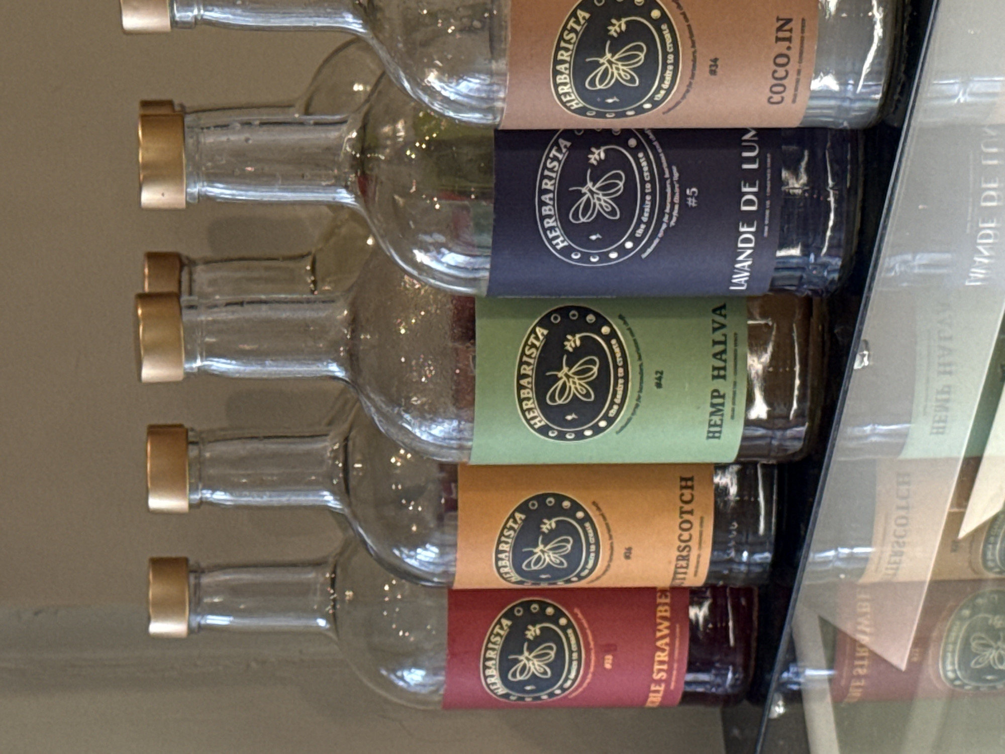 Six clear glass bottles with metallic gold-colored caps are displayed horizontally on a shelf in Moscow, Russia. The bottles are stacked vertically, with their caps facing left. Each bottle features a distinctively colored label wrapped around its midsection. The labels uniformly carry the brand name "HERBARISTA," accompanied by a circular logo depicting a bee or butterfly, and the slogan "the desire to create."
The visible labels from top to bottom are:
1. A brown label with the text "COCO.IN" and "#34".
2. A dark blue label with "LAVANDE DE LUM" (likely Lavande de Lune) and "#5".
3. A green label displaying "HEMP HALVA" and "#42".
4. An orange-brown label with "BUTTERSCOTCH" and "#26".
5. A red label showing "BLE STRAWBE" (likely Noble Strawberry) and "#23".
A sixth bottle with a red label is partially visible at the bottom.
To the right of the bottles, a reflective surface, possibly glass or a mirror, shows mirrored, inverted reflections of the labels and bottle necks. The background behind the bottles is a plain, light beige or tan wall. The display appears to be in an indoor setting, possibly a store, bar, or specialty food/drink establishment.