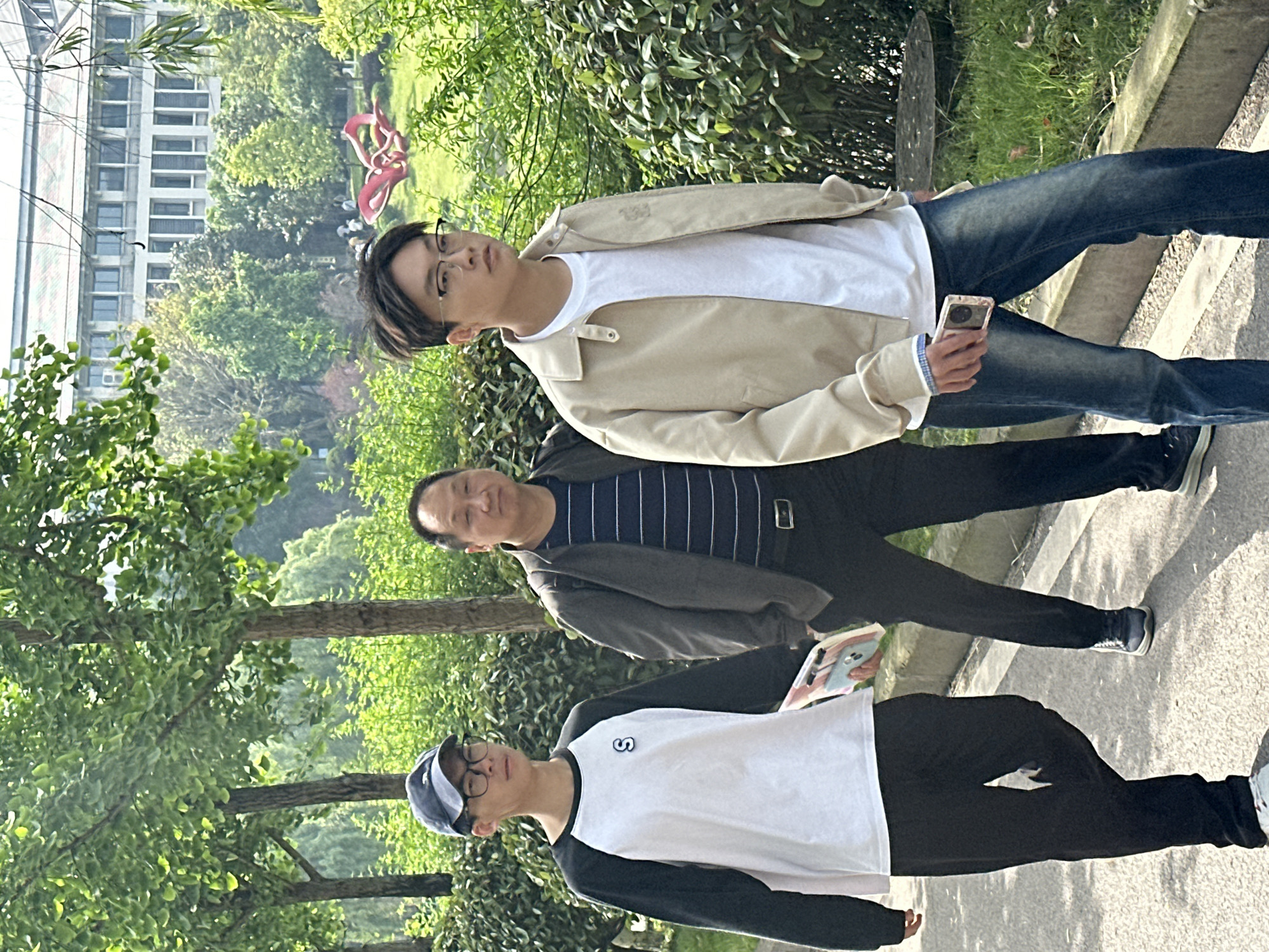 A daytime shot captures three individuals standing outside amidst a backdrop of trees and a building. The person in the front wears a baseball cap, glasses, a black and white shirt, and black pants. A middle-aged man in a jacket and striped shirt stands behind him. An individual wearing glasses, a white shirt, a tan jacket, and jeans holds a cell phone in the back. The scene is a park in Jiangjin, China.