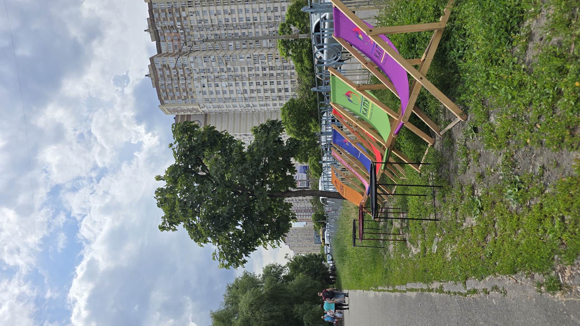 An outdoor urban scene in Kyiv, Ukraine, is captured under a partly cloudy sky. The foreground features a gray asphalt sidewalk on the left, occupied by four casually dressed adults walking. Two individuals walk away from the viewer, while two approach, partially obscured. To the right of the sidewalk, a grassy verge displays patchy green growth and bare earth. Six colorful wooden-framed lounge chairs are arranged along this verge, facing right. Their fabric seats are orange, pink, blue, red, green, and purple, respectively from left to right. The green and purple chairs clearly display Ukrainian text, "КАТПУ" and "Все буде добре," alongside a stylized graphic. In front of the leftmost chairs, three small, black-framed tables with black tops are present. A dark blue/gray metal fence with vertical bars separates the verge from a multi-lane asphalt road. Beyond the fence, several vehicles, including parked cars and a partially visible yellow vehicle, are on the road. A prominent deciduous tree with green foliage stands in the mid-ground, slightly right of center, while a line of dense green trees borders the left side of the sidewalk. In the background, tall, multi-story residential buildings with light-colored facades, numerous windows, and visible air conditioning units dominate the skyline, characteristic of Kyiv's residential architecture. Overhead utility lines are visible against the sky.
