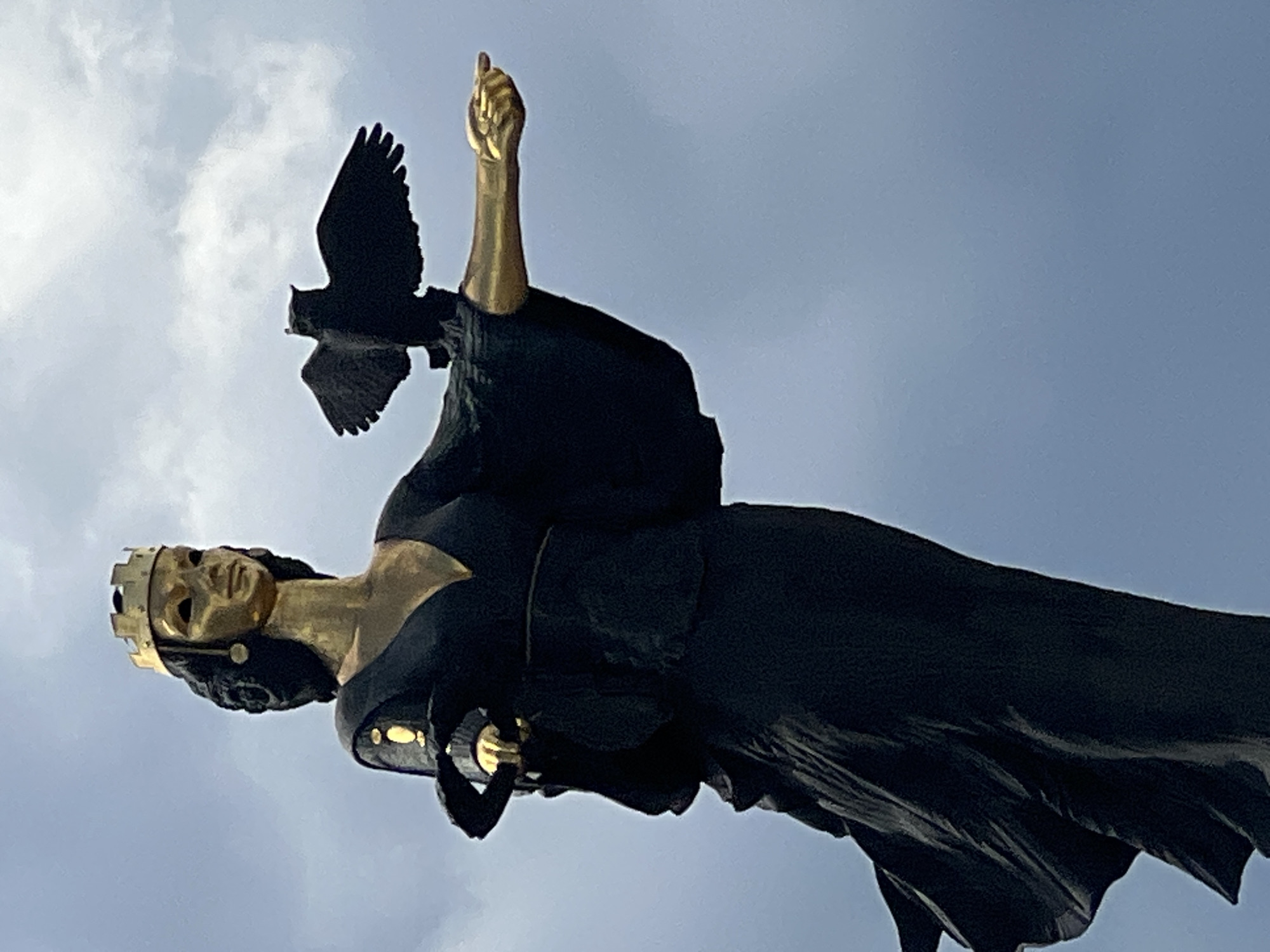 The image shows a close-up of the statue of the goddess Sophia in Sofia, Bulgaria. The statue is made of bronze and is gold-colored. The goddess wears a crown and holds a bird in her left hand, with her right hand extended forward. She is dressed in a long, flowing black robe. The statue is set against a backdrop of a cloudy, pale blue sky. The statue is a prominent landmark in Sofia, representing wisdom and knowledge.
