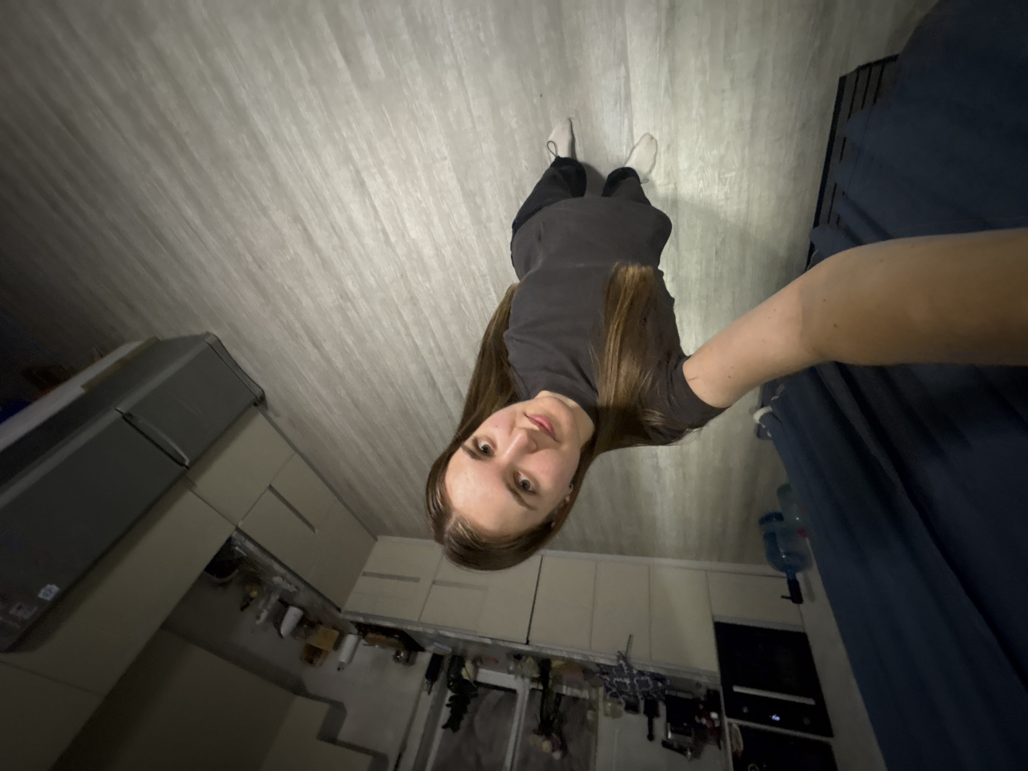 A young woman with long brown hair is standing in what appears to be a modern kitchen area. She is wearing a dark t-shirt and dark pants, with white sneakers on her feet. The kitchen features light-colored cabinetry and a grey countertop, with various items visible on the shelves, including bottles and what looks like kitchen appliances. The flooring is a light grey wood-grain laminate. The room is lit, suggesting it is daytime or that artificial lighting is in use. The camera angle is unusual, tilted and taken from a low perspective, making the woman appear to be standing upright while the surroundings are at an angle. The overall impression is a casual, indoor scene. The location is identified as being in the city of Perm, Russia.