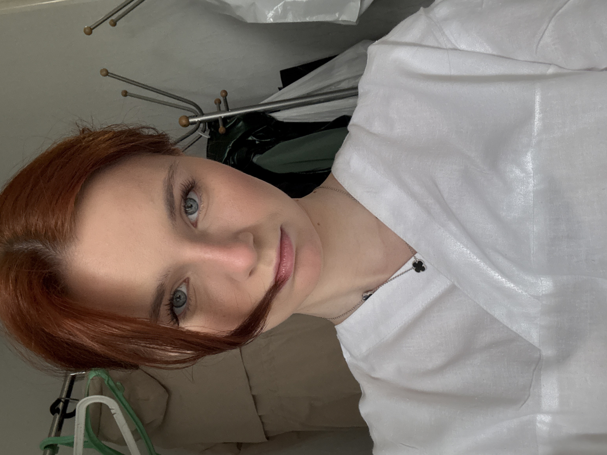 A close-up, portrait-style photograph shows a fair-skinned woman with red hair and blue eyes. She is wearing a white collared shirt. A silver necklace with a clover pendant is visible around her neck. The background is cluttered with a clothes rack, hangers, and various items of clothing. The photograph appears to be taken indoors, possibly in a closet or bedroom. There is no information to indicate this photo was taken in the city of Unknown, Russia.