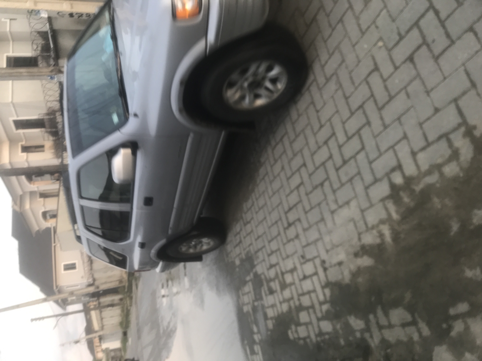 The image depicts an outdoor street scene in the Unknown, Unknown city, captured under soft, diffused daylight conditions, indicative of an overcast sky, early morning, or late afternoon.
In the foreground and midground, a silver-colored sport utility vehicle (SUV), consistent with an early 2000s Isuzu Rodeo or a mechanically similar model, is positioned on the right side of the frame. The vehicle's front is angled slightly towards the left, with the rear of the vehicle extending beyond the right frame edge. Visible features include silver paint, black fender flares over the wheel wells, a roof rack, and multi-spoke alloy wheels. The front right headlight assembly is partially visible. The windshield shows reflections and indistinct forms within the vehicle's interior, possibly a sunshade or objects on the dashboard. Motion blur is evident on the vehicle's wheels, suggesting either slow movement of the vehicle or camera shake during capture.
The ground surface beneath the vehicle and extending to the left consists of interlocking grey pavers laid in a herringbone pattern. Irregular patches of standing water or wetness are visible on the left half of the paved area, suggesting recent precipitation or accumulated surface water.
In the background, a residential or mixed-use urban environment is visible. A tall, slender utility pole stands vertically on the far left. Several buildings are present, primarily light-colored structures with dark roofs. The leftmost visible building is a single-story structure. Adjacent to it, a larger, multi-story building with light-colored walls, a dark gabled roof, multiple windows, and decorative architectural elements is prominent. A fence, topped with multiple strands of what appears to be coiled barbed or razor wire, runs in front of the rightmost visible building, which shares a similar architectural style to the adjacent larger structure. No individuals are visible in the scene.