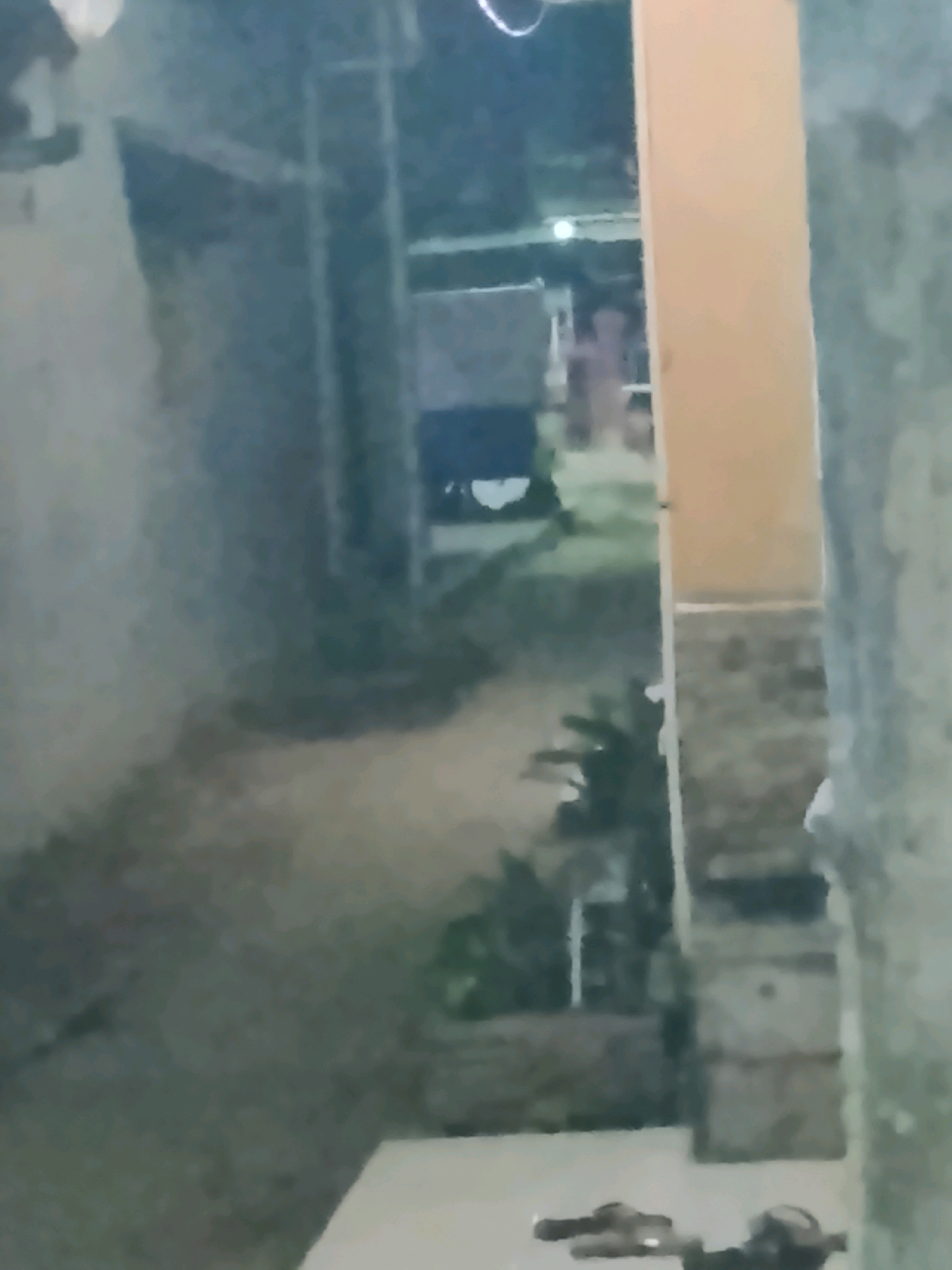 The image depicts a dimly lit alleyway or pathway between buildings. On the right side of the frame, a light-colored pillar is prominent, with textured walls on either side. The ground appears to be paved or concrete. In the background, there are glimpses of structures, possibly houses or other buildings, with some indistinct shapes that could be laundry hanging. The overall scene is blurry and lacks clear details due to low light conditions and potential motion blur. The location is identified as Ujungrusi, Indonesia. The bottom of the image shows two pairs of footwear.