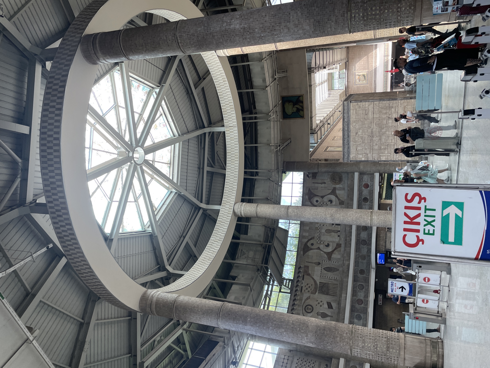 The image shows the interior of a public transportation hub, likely a metro station, in Istanbul, Turkey. The architecture features a large, circular skylight with a metal frame, supported by thick, tiled pillars. A prominent sign in Turkish and English reads "ÇIKIŞ EXIT" with a green arrow pointing to the right, indicating an exit. Several people are visible, some walking, others standing near a ticket barrier or waiting. One sign indicates "METRO" with an arrow. The overall environment appears to be a modern, well-lit interior with a tiled floor and decorative wall art. The atmosphere seems to be one of a busy transit point with people moving through.