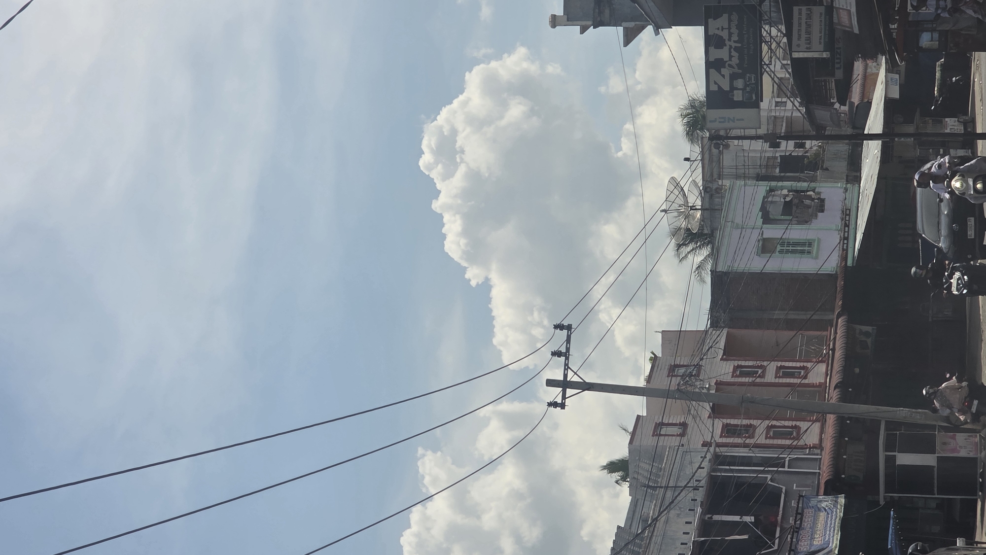 The image captures a street scene in Teungoh Baro, Indonesia, under a partly cloudy sky. Buildings line the street, displaying diverse architectural styles and signage, including commercial advertisements. Overhead power lines crisscross the view, connected to utility poles. The presence of satellite dishes indicates access to broadcast television services. Motorbikes with riders are visible on the street, suggesting the area is moderately active with vehicular traffic. The lighting indicates daylight hours. The composition of the shot is angled upwards, prioritizing the sky and clouds relative to the buildings and street below.