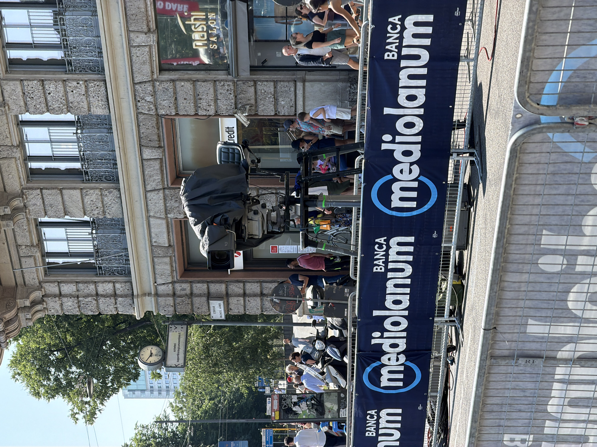 Here is a detailed description of the image:
The image captures a street scene in Milan, Italy, prominently featuring the banner of Banca Mediolanum along a street, likely Corso Buenos Aires, as suggested by the street sign. The image is taken from a slightly elevated angle.
The scene is filled with people, some of whom are standing behind the barriers that are adorned with the Banca Mediolanum banner. A covered camera is set up, possibly for filming an event or providing coverage.
Street furniture is noticeable: street signs, traffic lights, a clock attached to a pole indicating "scriveteci support@orologicomunemilang.it," and the name of the street.
The architectural background consists of a building with many windows and decorative ironwork. Several signs are visible, "Credit" and "Nashi Salon."
The overall setting conveys a public space in a busy urban environment with indications of possible street events.