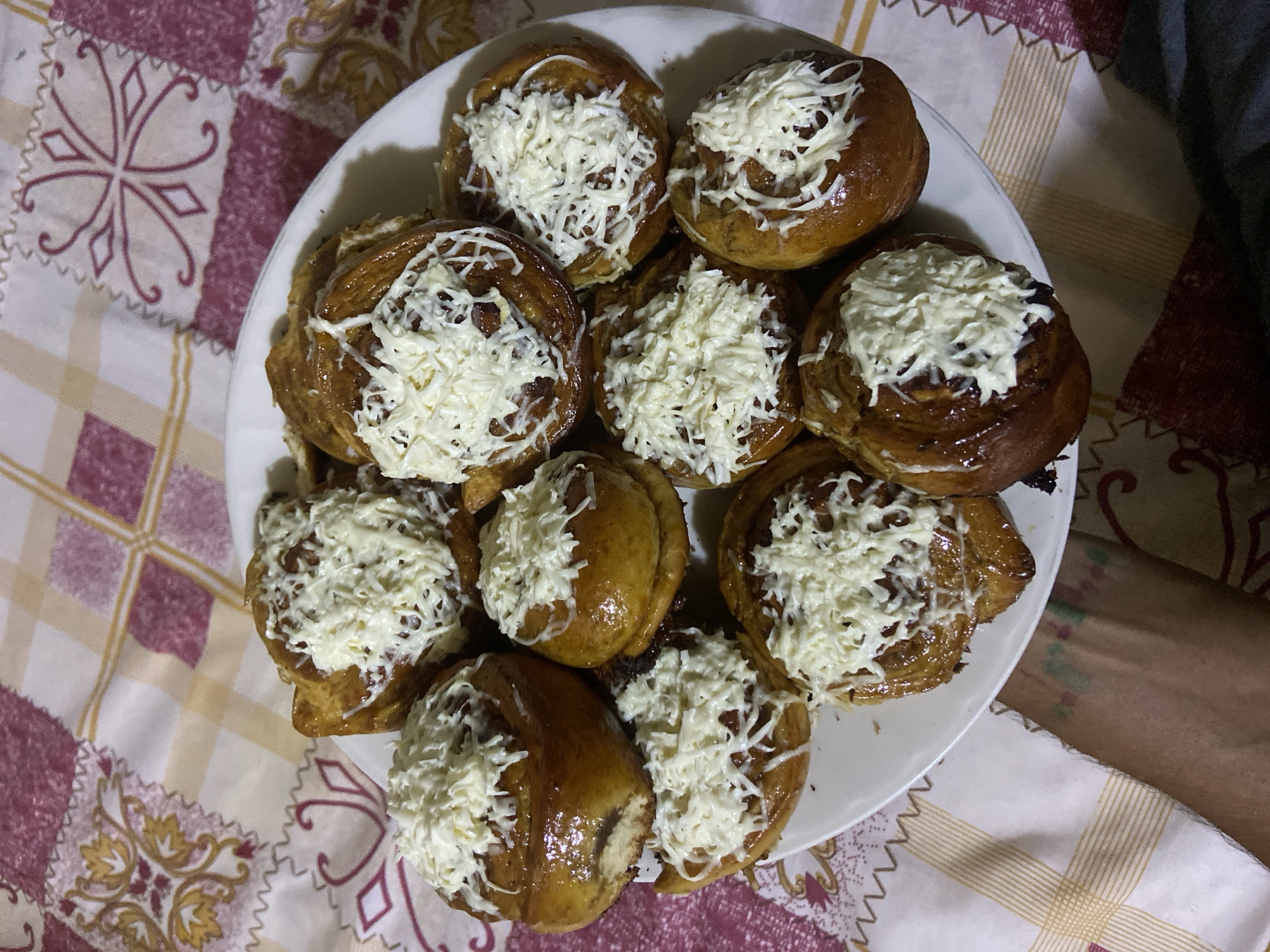 A plate of cinnamon rolls topped with grated cheese is presented on a table covered with a patterned tablecloth. The cinnamon rolls are golden brown and appear glazed. The tablecloth has a repeating geometric pattern in shades of maroon and beige. A person's arm, with tattoos, is visible in the lower left corner of the image, resting on the table. The scene is indoors and appears to be a casual setting. There is no specific information about the location in the city of Los Velásquez, Venezuela within the image itself.