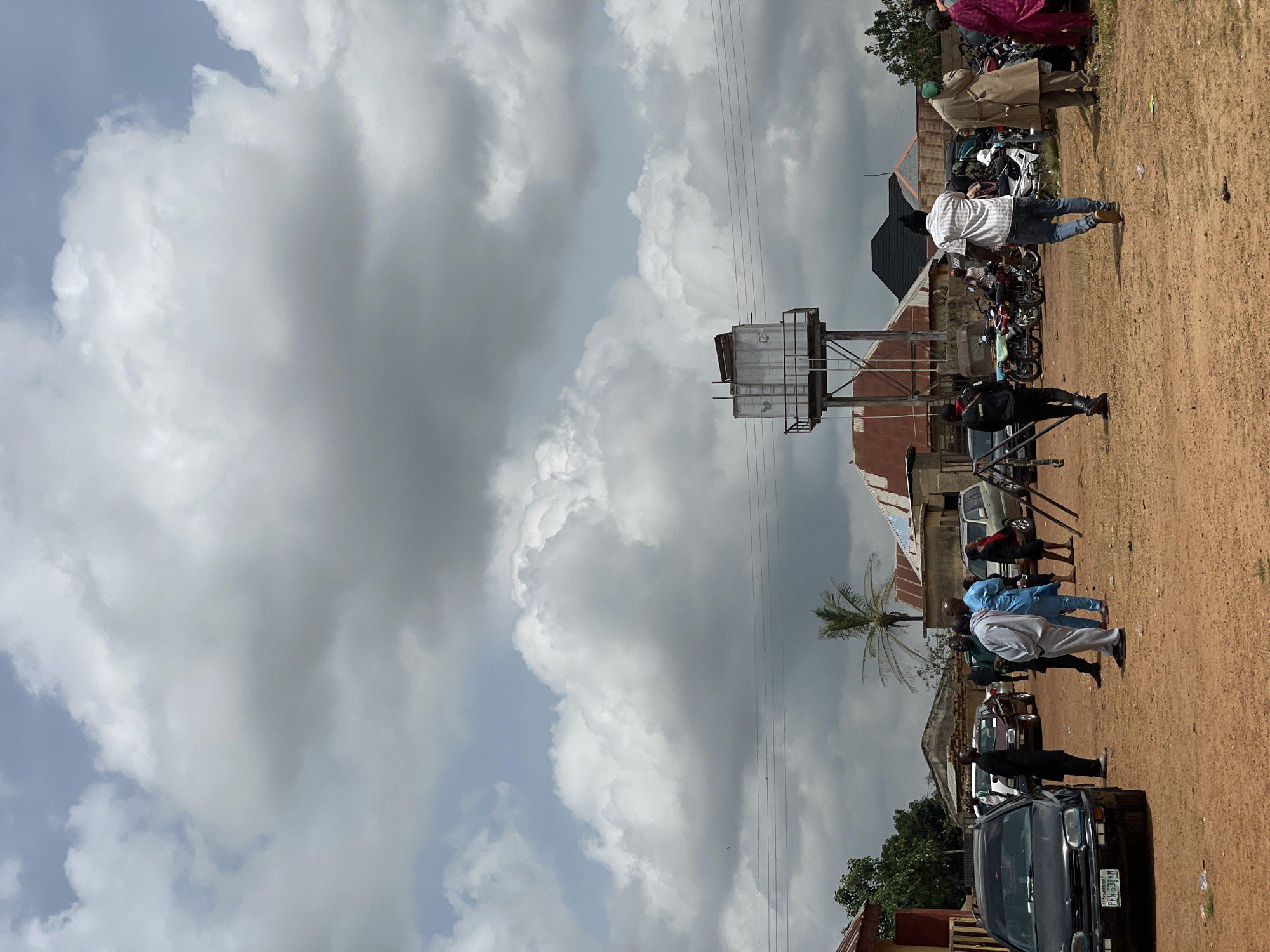 In Kassa, Nigeria, under a cloudy sky, a group of people are gathered in an open, dirt lot. Several vehicles are parked, including a black sedan in the foreground with a license plate that reads "PKN 631 KM". To the right of the sedan, a group of men are engaged in conversation, some dressed in traditional attire. Further back, a water tower stands tall, overlooking the scene. Motorcycles are also visible, parked together, suggesting a mode of transportation for some of the attendees. The background reveals residential buildings with corrugated metal roofs, indicative of the local architecture. The overall atmosphere appears to be a community gathering or event.