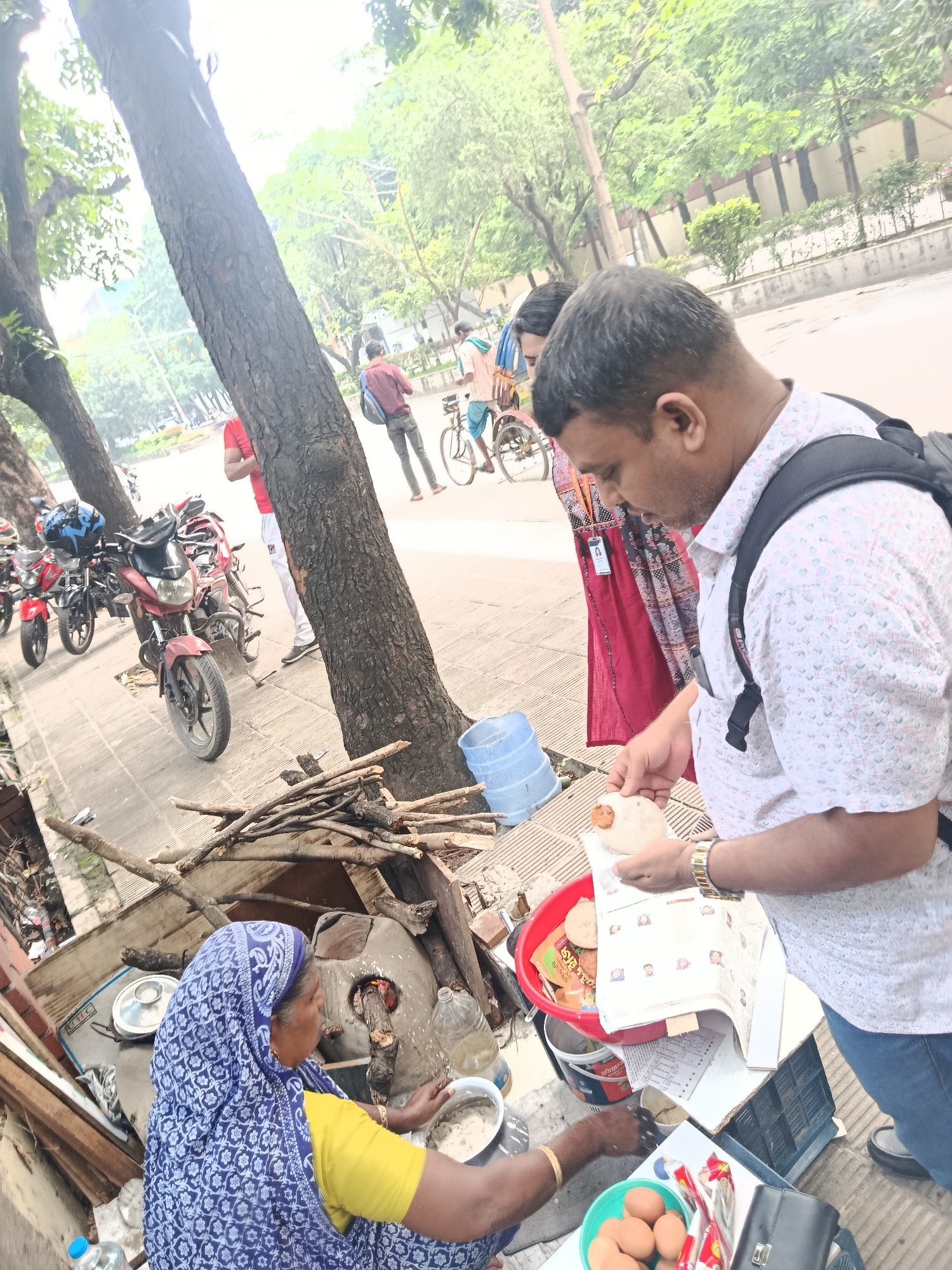The image shows a street food vendor in Dhaka, Bangladesh. An older woman wearing a blue patterned headscarf and yellow top is cooking food on a traditional wood-fired stove. She is seated and working with ingredients in a bowl.
A man with a backpack is standing next to her, examining a flatbread-like item in his hands while holding a book. There's a red bowl with more flatbreads and other items on a small table or crate. A container of eggs is also visible.
Behind the vendor, there's a pile of wood for the stove. In the background, there are several motorcycles parked on the sidewalk, and people are walking or riding bicycles on the street. Trees line the street, suggesting an urban environment.