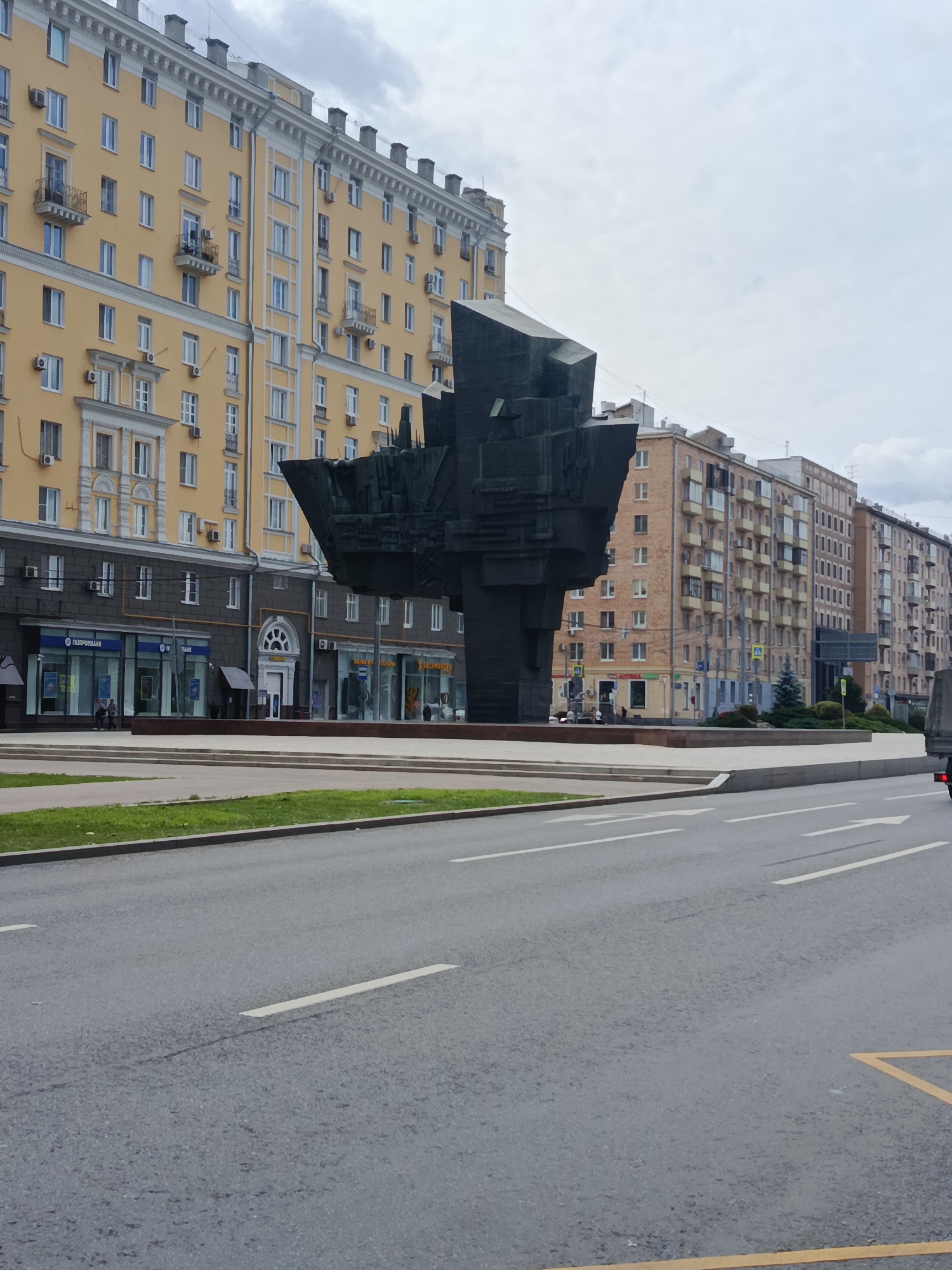 The image depicts a wide street in Moscow, Russia, featuring a large, abstract black sculpture in the foreground, a yellow apartment building to the left, and a series of brick apartment buildings to the right. The sculpture is a prominent, angular, dark metal structure with multiple geometric planes. The yellow building, typical of Stalinist architecture, has numerous windows and decorative cornices. The brick buildings on the right are also multi-story apartment blocks with balconies. The street is paved, with visible lane markings and a yellow "no parking" zone on the right side. There are some pedestrians visible near the building entrances and a few cars on the street. The sky is overcast, suggesting a cloudy day. The overall scene captures an urban environment with a mix of monumental architecture and everyday city life.