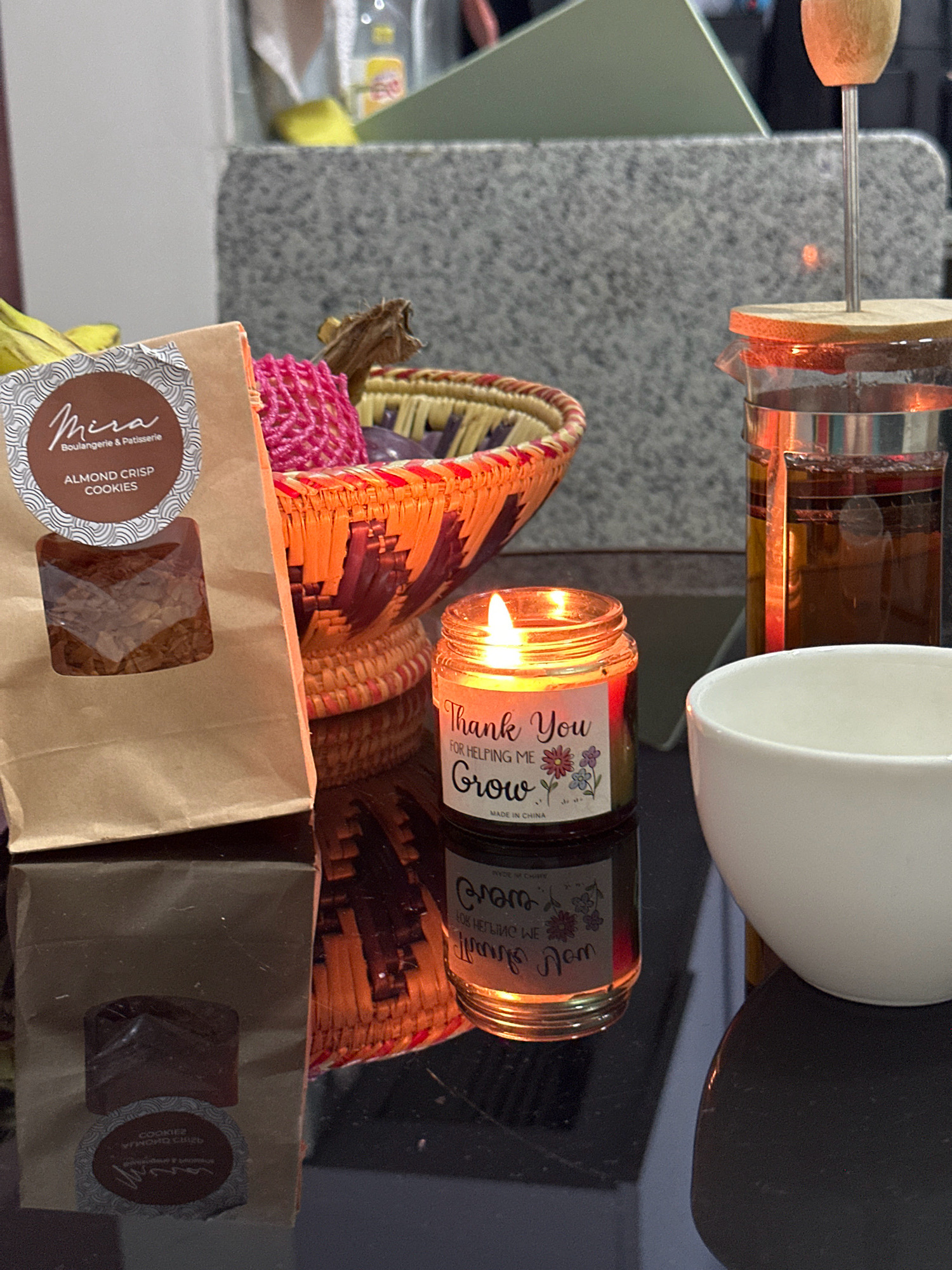 The image depicts a close-up view of a table setting, likely on a reflective black surface. In the foreground, a small, lit candle in a glass jar glows warmly, with text on the label reading "Thank You For Helping Me Grow." To the right of the candle is a white ceramic bowl. Behind the candle and bowl, a glass French press filled with amber-colored liquid is visible, suggesting tea or coffee. To the left, a brown paper bag with a clear window reveals almond crisp cookies, with a circular label bearing the text "Mira Boulangerie & Patisserie." A woven basket with colorful threads and dried plant material is situated behind the cookie bag. In the background, a mottled gray surface and a hint of greenery are visible. The overall impression is one of a cozy, possibly domestic, setting. No specific location in Lalitpur, Nepal, can be determined from the image.