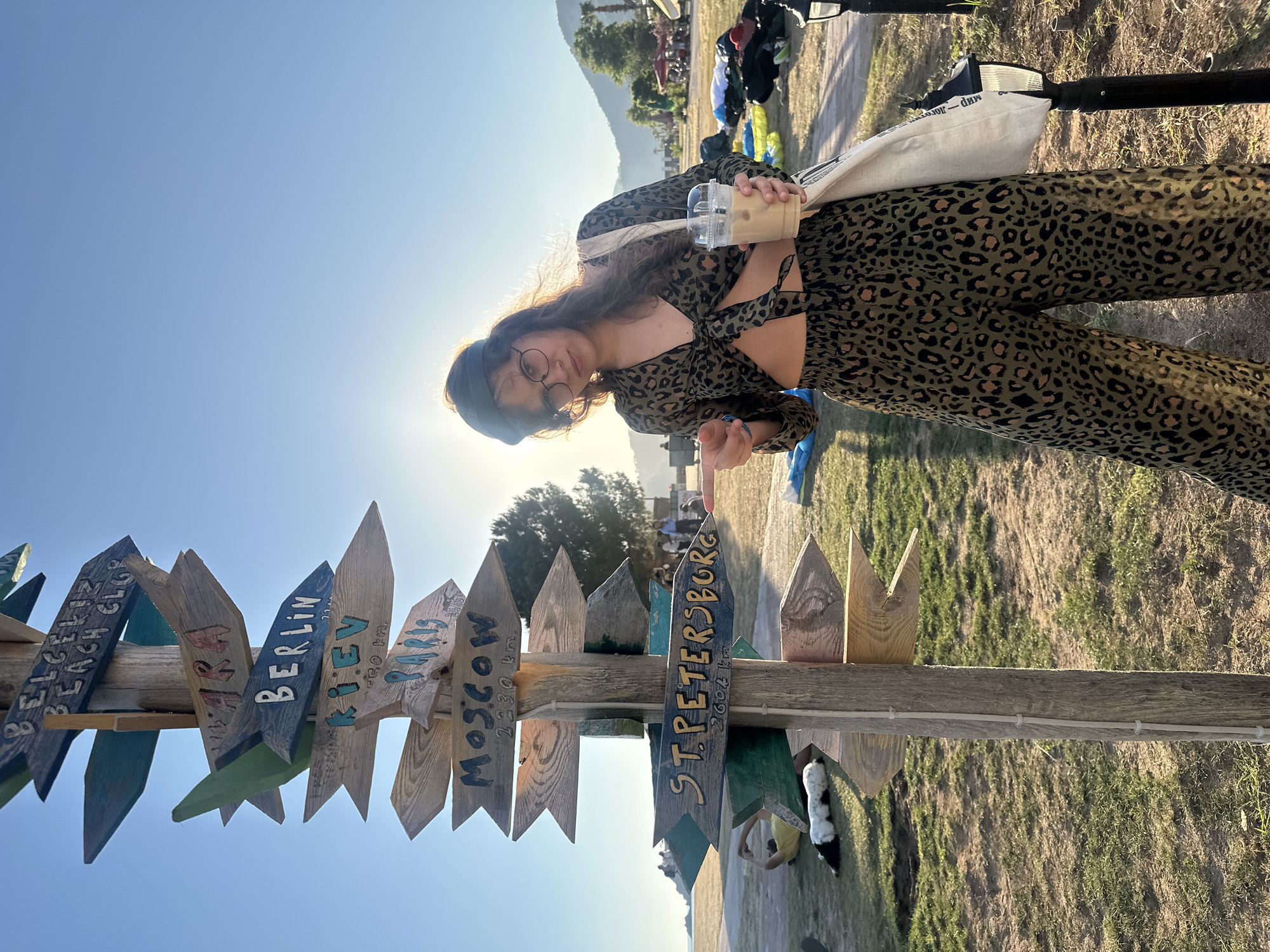 A young woman in a leopard-print outfit stands next to a signpost in Fethiye, Turkey. She holds a plastic cup with a beverage, and the sun shines behind her. The signpost is made of wood and has arrows pointing in different directions, each with the name of a city: "Beach Club," "Berlin," "Kiev," "Paris," "Moscow 2230 km," and "St. Petersburg 2602 km." The background includes grass, trees, a mountain range, and other people in the distance. A white tote bag hangs from her shoulder.