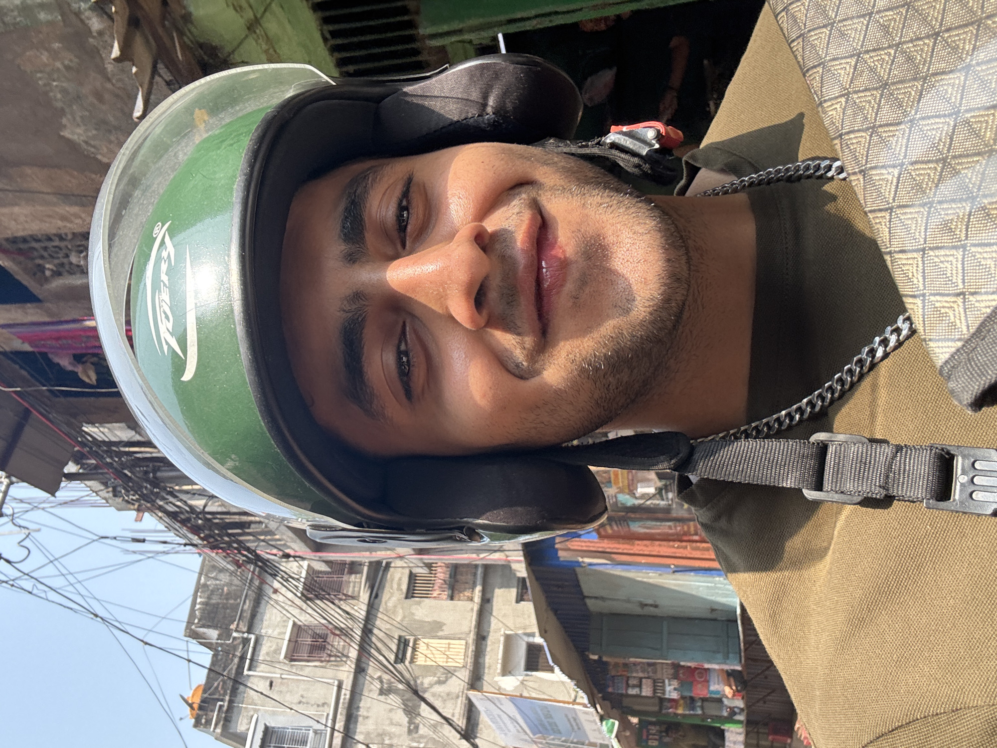 A close-up selfie shows a young man wearing a green and black helmet. He appears to be on a motorcycle, as the helmet suggests. He has a light beard and dark hair, and a chain necklace is visible. The background reveals an urban street scene with buildings and tangled overhead wires, typical of Kolkata, India. Buildings are worn and have multiple small windows, suggesting residential or mixed-use structures. Various commercial signs and possibly storefronts are visible in the background. The lighting suggests it is daytime, potentially mid-morning or afternoon.