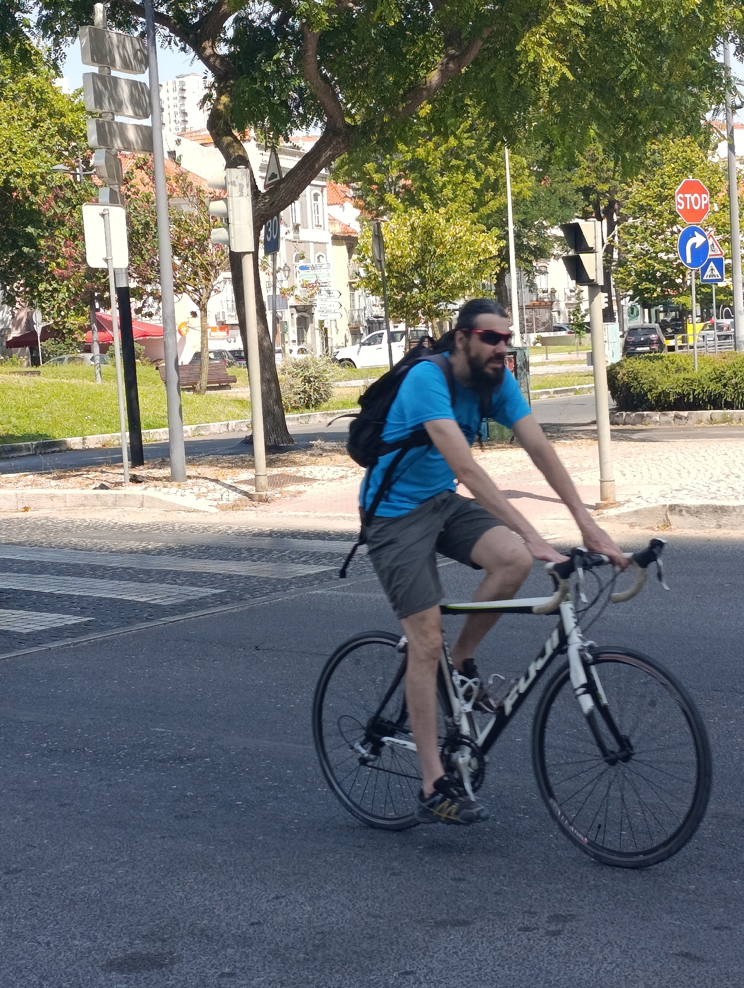 A bearded man wearing sunglasses, a blue t-shirt, and grey shorts rides a bicycle on an asphalt road in Setúbal, Portugal. He has a black backpack on. The road has a pedestrian crossing. In the background, there are trees, buildings, traffic lights, and street signs. The man is in motion, with his hands on the handlebars and his legs pedaling. The sun is shining.